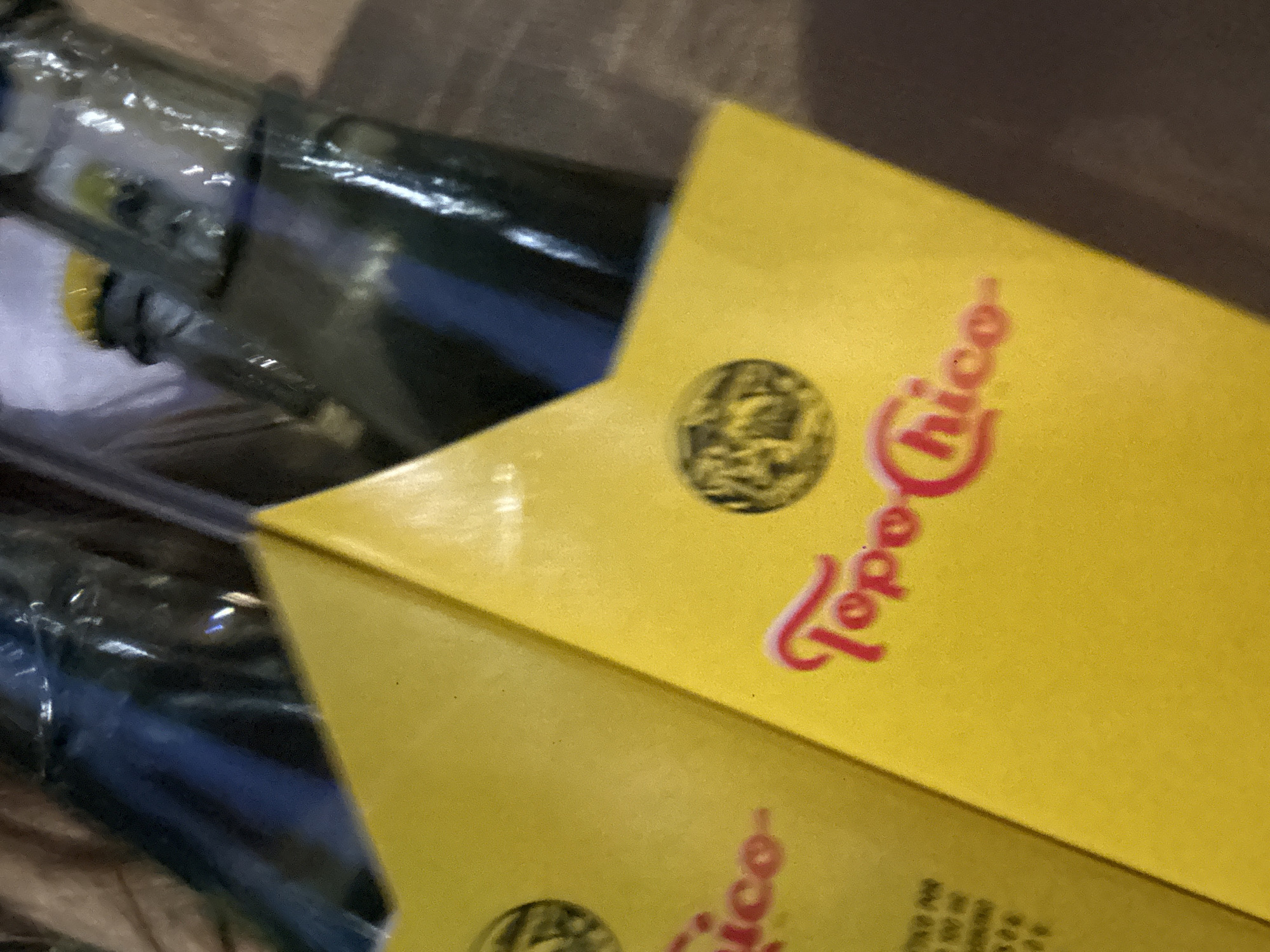 A close-up shot shows bottles of Topo Chico mineral water in their yellow cardboard packaging. The logo and name "Topo Chico" are prominently displayed in red on the packaging. The bottles appear to be made of glass, with moisture visible on their surfaces. The scene's location is not discernible from the image.