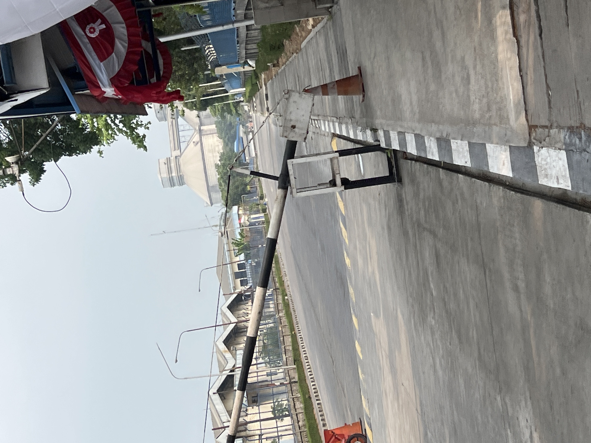 The image depicts a street scene in Klapanunggal, Indonesia, with a focus on a security barrier and industrial buildings in the background. A black and white striped barrier arm is extended across the road, suggesting a checkpoint or entrance. An orange traffic cone is visible on the sidewalk to the right of the barrier. In the distance, several large, industrial-style buildings with conical roofs are present, characteristic of a factory or processing plant. The sky is overcast, and trees are visible along the side of the road. A decorative red and white fabric banner hangs from an overhang on the right side of the frame. The overall impression is that of an industrial zone or facility in a semi-urban environment.
