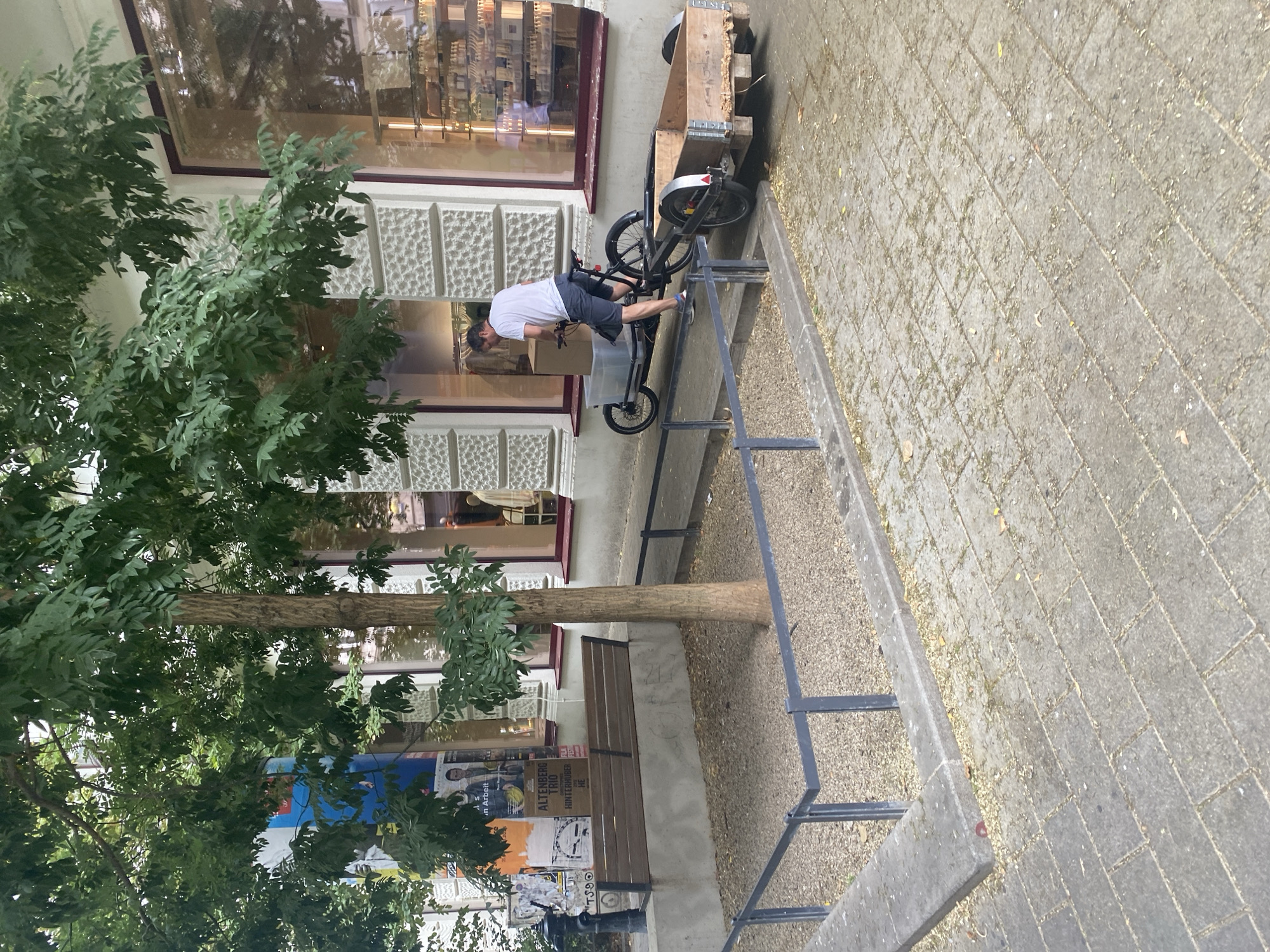 A man in a white t-shirt and dark shorts is riding a bicycle with a long trailer attached. He is moving along a paved sidewalk next to a building with large windows and an ornate facade. The trailer is wooden and appears to be empty. The bicycle is black and has a large cargo area in front of the handlebars, which contains a clear plastic container and a cardboard box. A tree with green leaves shades a portion of the scene. In the background, a park bench is visible next to a tree-lined area with gravel. The image appears to have been taken in an urban setting, possibly on a street in Vienna, Austria, given the architectural style of the building and the presence of posters on the wall.
