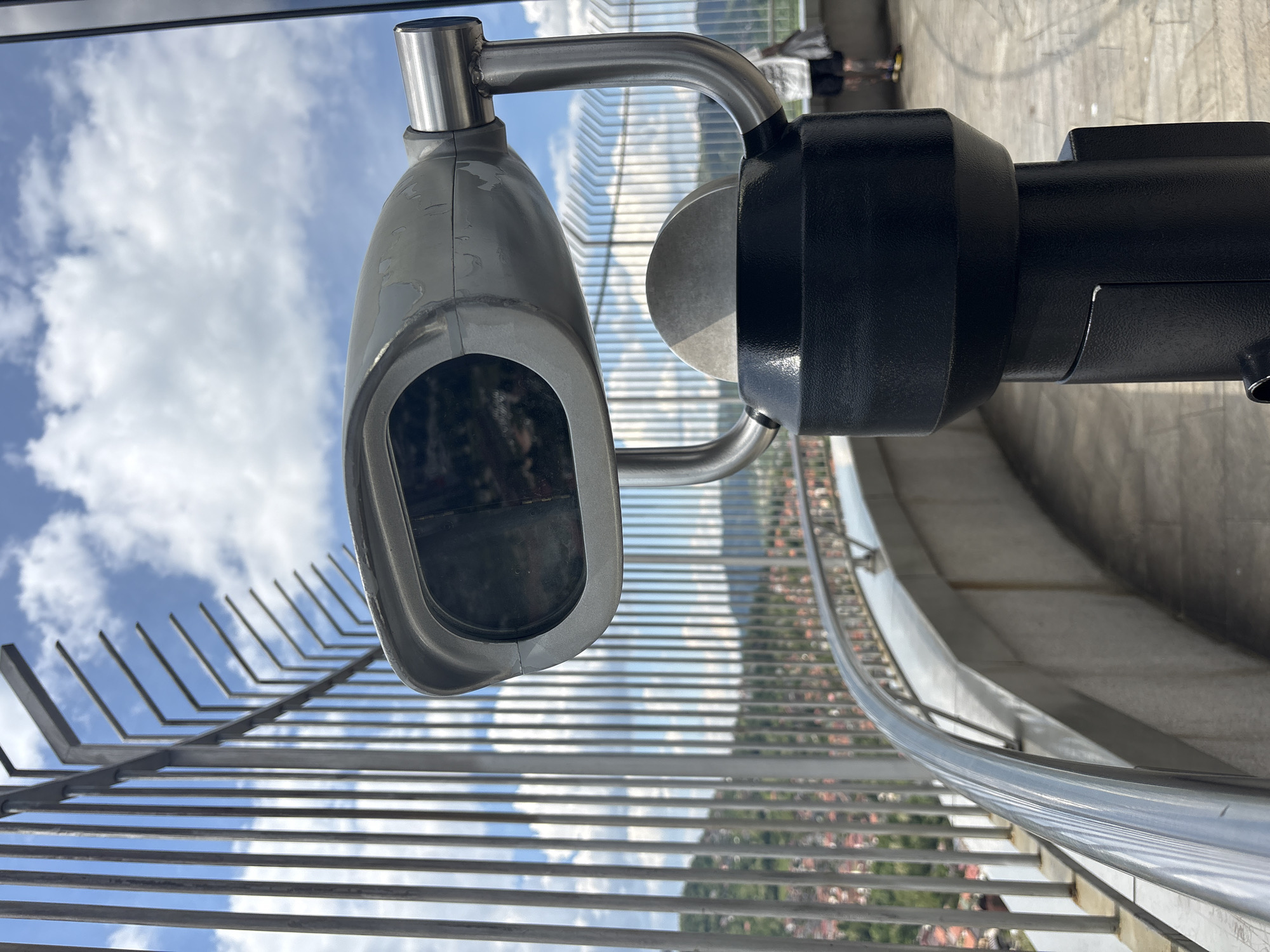 The image depicts a coin-operated telescope mounted on a railing, likely at an observation point overlooking the city of Sarajevo, Bosnia and Herzegovina. The telescope is silver and black. The railing is metal with horizontal bars. The background shows a cityscape and landscape under a partly cloudy sky.