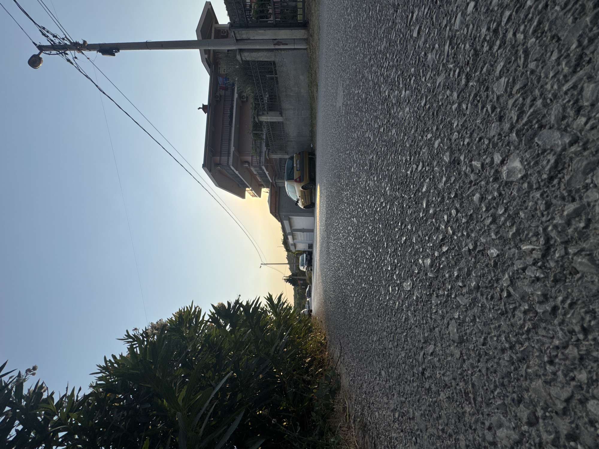 The image depicts a street scene in Sutomore, Montenegro, during the golden hour. The perspective is low to the ground, focusing on the asphalt road. To the left, a dense green bush with some white flowers lines the road, partially obscuring the view further down the street.
The road itself is paved with dark grey asphalt, textured with visible stones and gravel. Sunlight casts a warm glow, particularly near the horizon. In the middle ground, several cars are parked or driving on the road. A yellow hatchback is prominently parked on the right side of the street.
Further down the road, houses and buildings are visible. On the right, a multi-story house with balconies and a dark metal fence is situated. A concrete utility pole with a street lamp and a tangle of electrical wires stands beside it. Some garages or sheds with white doors are also present. The sky is a clear, pale blue, transitioning to a lighter yellow-orange hue near the setting sun on the horizon. The overall atmosphere is tranquil and serene.