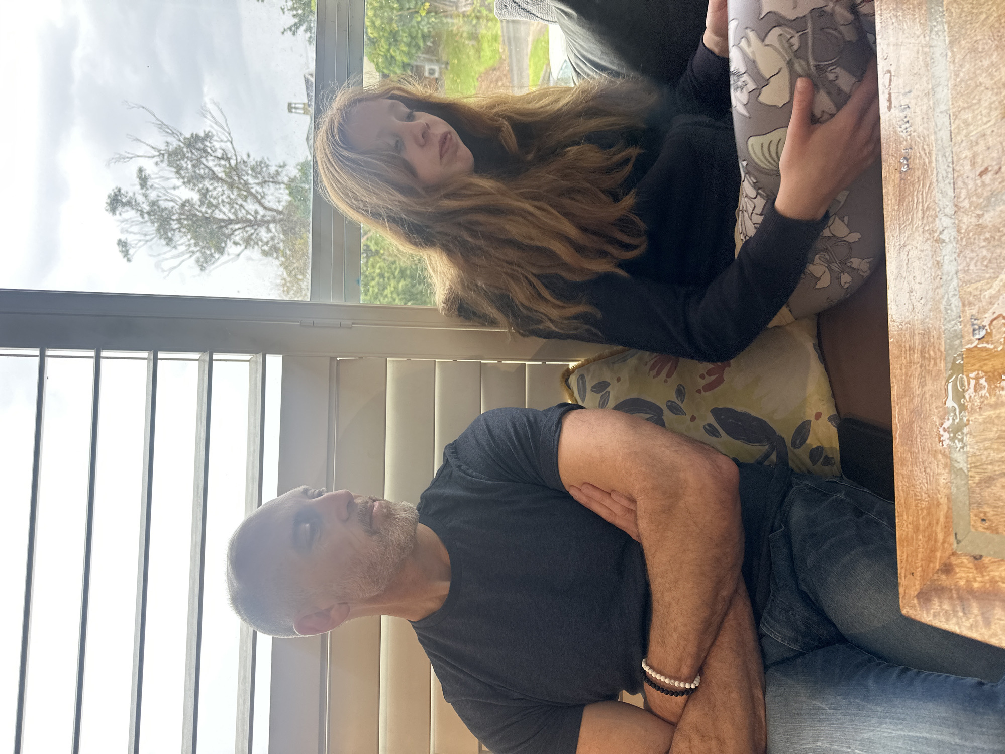 In Nambucca Heads, Australia, a man and a young woman are seated indoors, facing away from each other. The man, with short gray hair and a beard, is wearing a dark blue t-shirt and blue jeans. His arms are crossed, and he is looking to his left. The woman, with long auburn hair, is wearing a black top and patterned leggings. She is looking down. They are seated on a couch with a patterned cushion between them. Behind them is a window with blinds, and outside the window, a tree and a cloudy sky are visible. In the foreground, a wooden table with a light-colored surface is present.