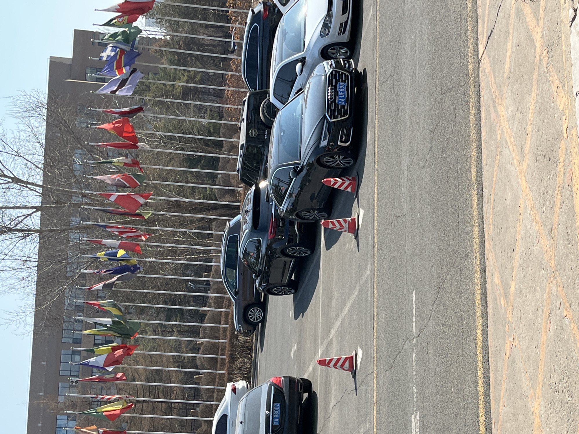 The image shows a parking lot in Changchun City, China. Several cars are parked, demarcated by yellow painted lines and traffic cones. A building stands in the background, adorned with numerous flags from various countries. Bare trees are visible to the left of the building. The sky is clear and blue.