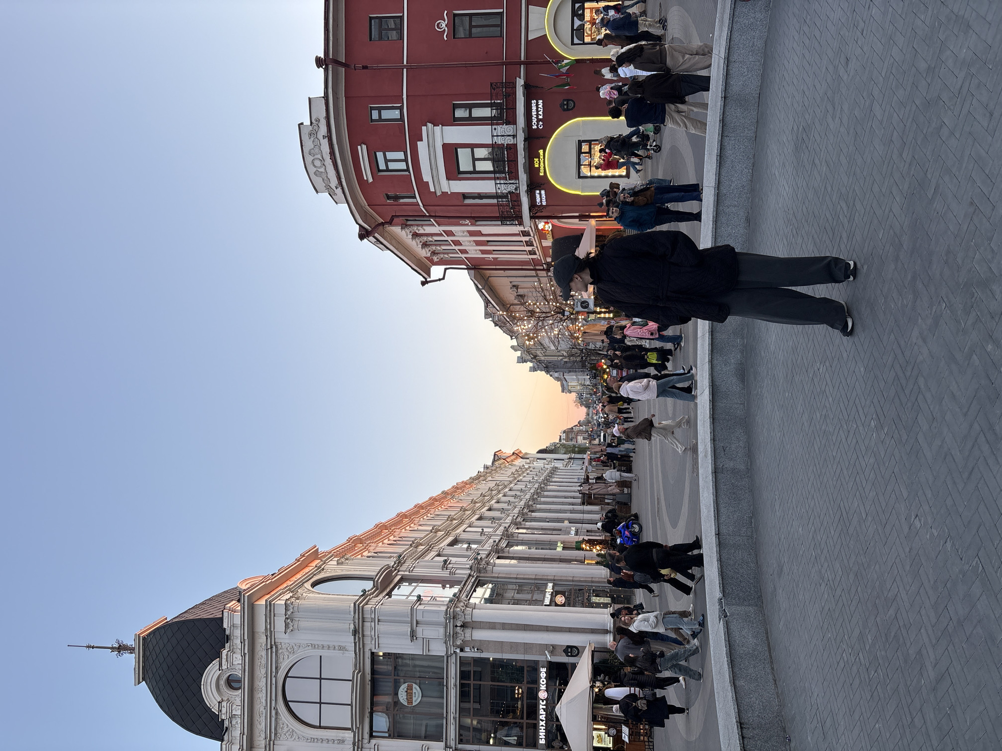 The image captures a bustling street scene in Kazan, Russia, likely on Bauman Street. It showcases a mix of pedestrians strolling along the cobblestone street. The buildings on either side of the street display diverse architectural styles, with a red building featuring a rounded corner and a lighter-colored building with columns. The scene is well-lit, suggesting daylight conditions. The presence of numerous individuals and businesses implies a lively and active environment.