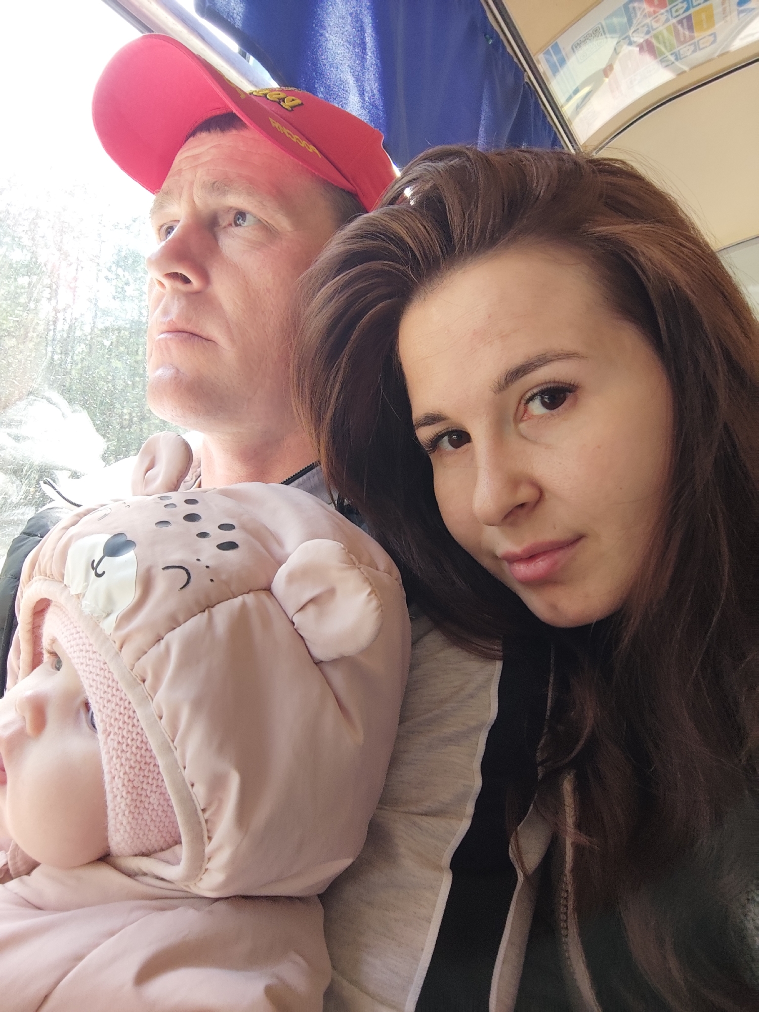 The image depicts a family—an adult male, an adult female, and an infant—traveling inside what appears to be a public transport vehicle, likely a bus or train. The setting is Garevaya, Russia, during the daytime, indicated by the bright light streaming through the window.
The adult male, on the left, wears a red baseball cap with yellow text (possibly "REESE'S") and a dark jacket. He looks out the window with a serious, contemplative expression. His gaze is directed upwards and outwards.
Beside him, on the right, the adult female with long brown hair looks towards the camera with a calm, neutral, or slightly smiling expression. She is wearing a light-colored top with a distinctive black and white vertical stripe.
In front of the female, the infant is bundled in a warm, pink padded hooded overall designed with a bear face, complete with small ears, black dots for eyes and nose, and curved lines for a mouth. A light pink knitted hood is visible underneath. The baby, like the male, is also looking out the window, appearing curious.
The interior of the vehicle includes a prominent window frame, blue fabric along the upper ceiling, and a light-colored, curved ceiling section. A diagram or map with numbers (e.g., "1," "2") and symbols is visible on the ceiling. The view outside the window is bright but blurred, suggesting motion. The overall impression is a quiet moment of travel and observation within the family unit.