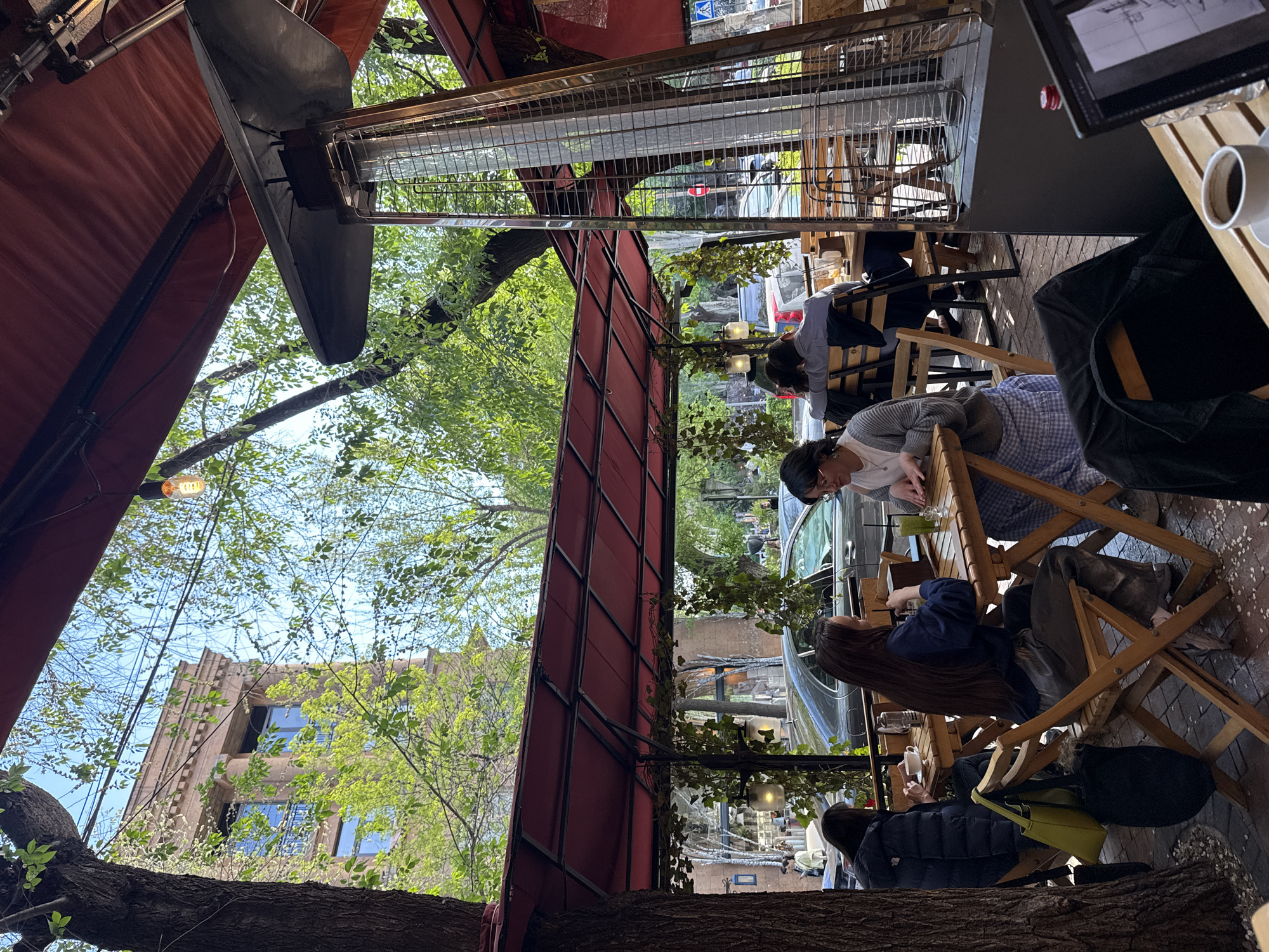 The image depicts an outdoor cafe setting in Yerevan, Armenia. Several individuals are seated at wooden tables and chairs, some engaged in conversation. The scene is partially covered by a red awning and outdoor heaters. A multi-story building is visible through trees. A car is parked on the street adjacent to the cafe. A coffee cup and electronic tablet are on one of the tables.