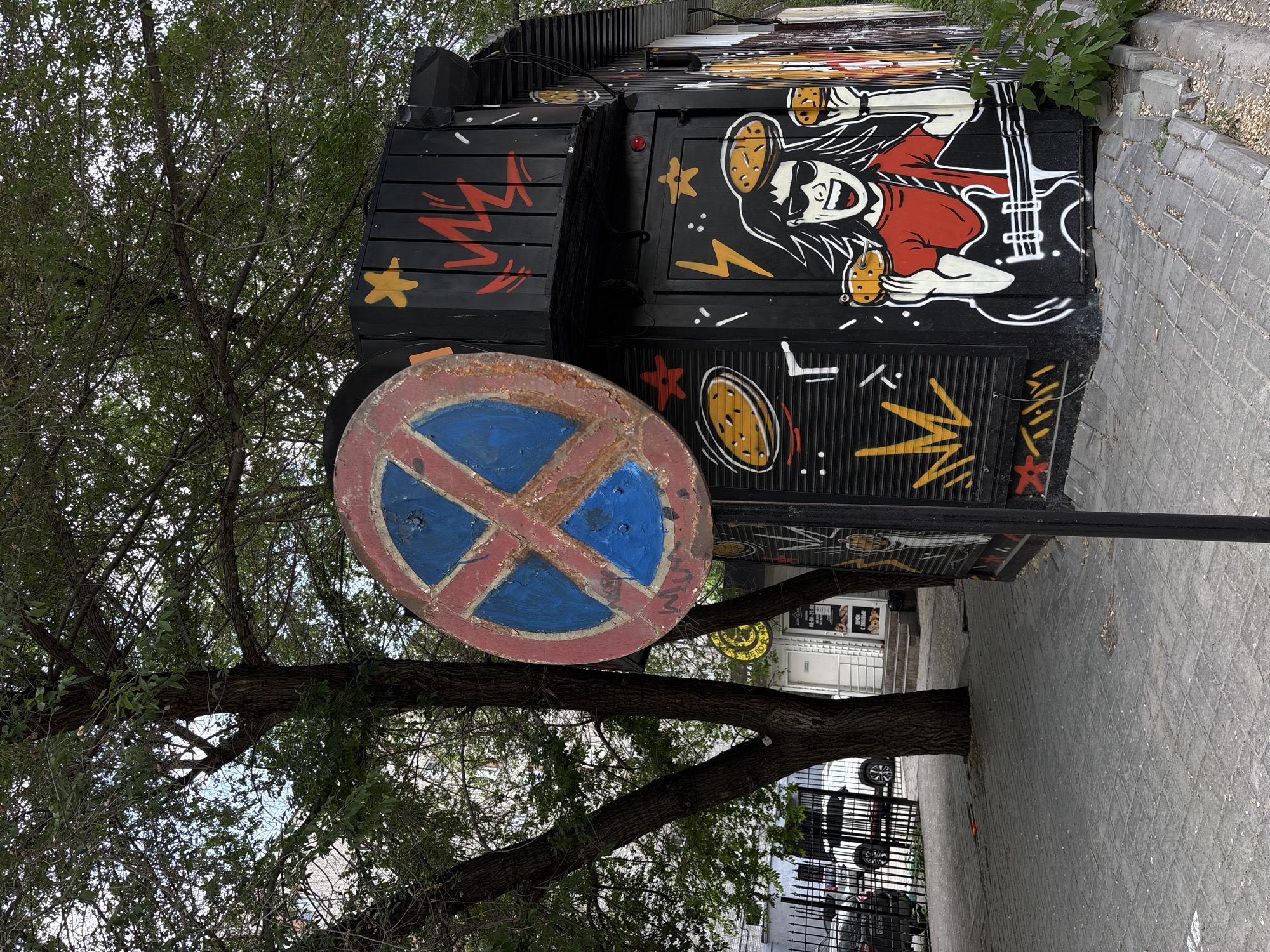 An outdoor shot captures a street scene in Novosibirsk, Russia. In the foreground, a weathered, round "No Parking" sign with a red border and a blue 'X' is positioned on a metal pole. To the right of the sign, a black kiosk or small building is adorned with vibrant graffiti. The artwork depicts a cartoon character with sunglasses and wild hair, wearing a red shirt and playing a guitar, while holding two burgers. Decorative yellow stars and red lightning bolts are also painted on the building's exterior. Behind the kiosk, a tree with dense green foliage provides shade. In the background, a paved walkway leads to a building entrance with stairs, and a metal fence encloses a street with parked cars, suggesting an urban setting.
