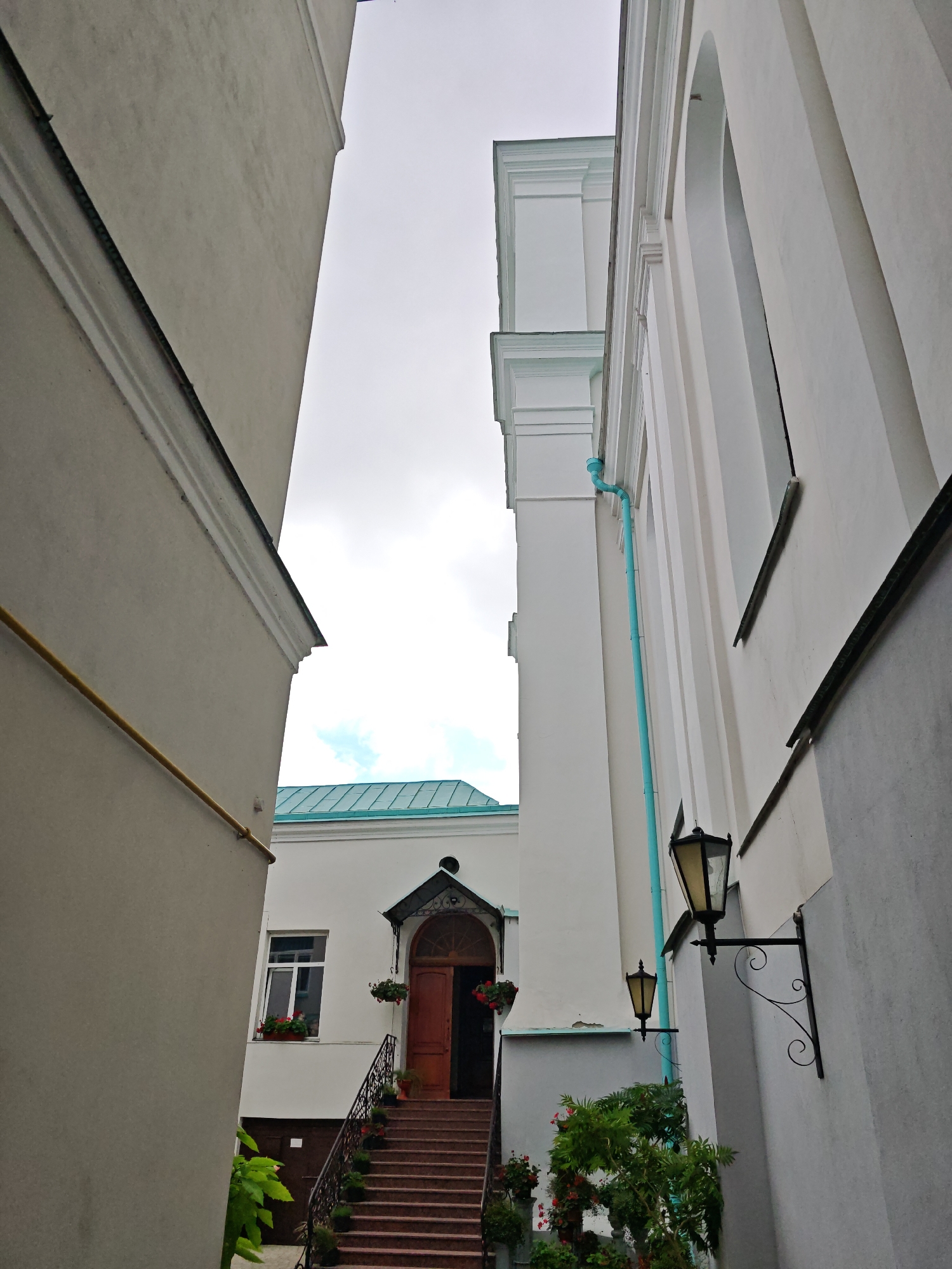 The image depicts a narrow alleyway between two buildings, likely in the city of Hrodna, Belarus. The building on the left is a light gray facade. The building on the right is white with architectural details, including pilasters and arched windows. A bright teal downspout runs vertically along the white building. Two wrought-iron wall lamps with yellow glass shades are mounted on the white facade. In the background, a set of red-brown stairs leads up to a wooden door under a decorative canopy. A window with red flowers in a planter is visible to the left of the door. Green foliage and potted plants are present near the base of the stairs and on the right side of the alley. The sky is overcast.