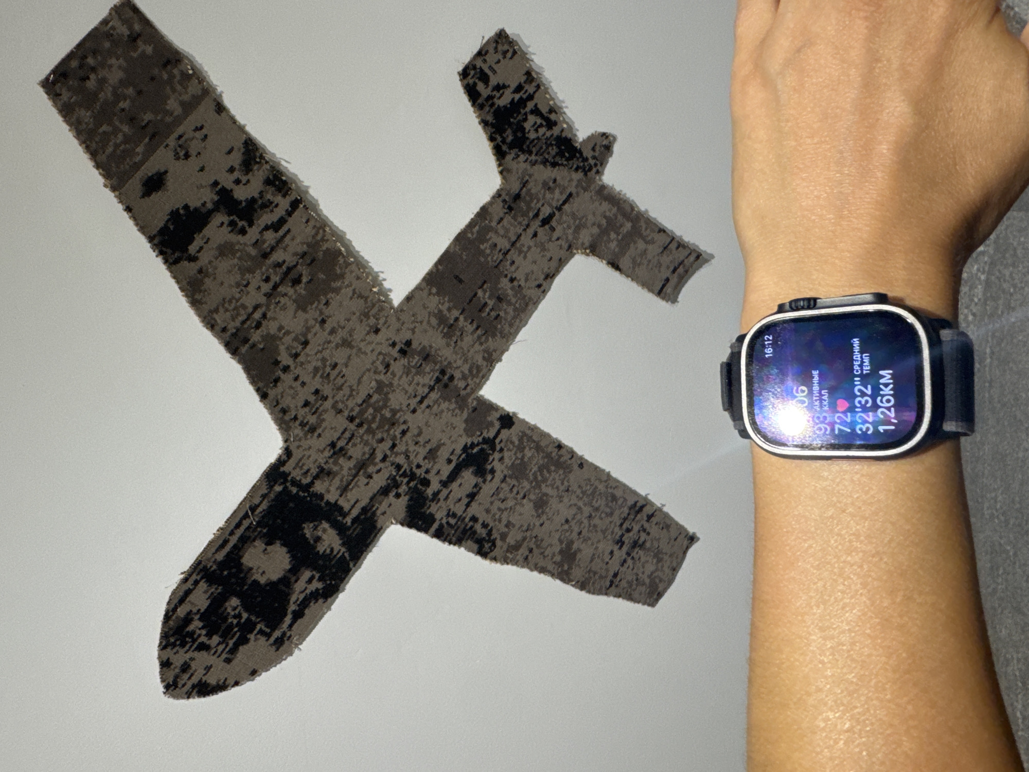The image shows a person's left forearm and wrist wearing a smartwatch with a dark strap. The smartwatch displays information including the time "16:12", "06", "93 ккал", "72", "32'32''", and "1,26км". In the background, on a light-colored wall, is a camouflage-patterned cutout of an airplane. The location is the city of Arkhangel’skoye, Russia.