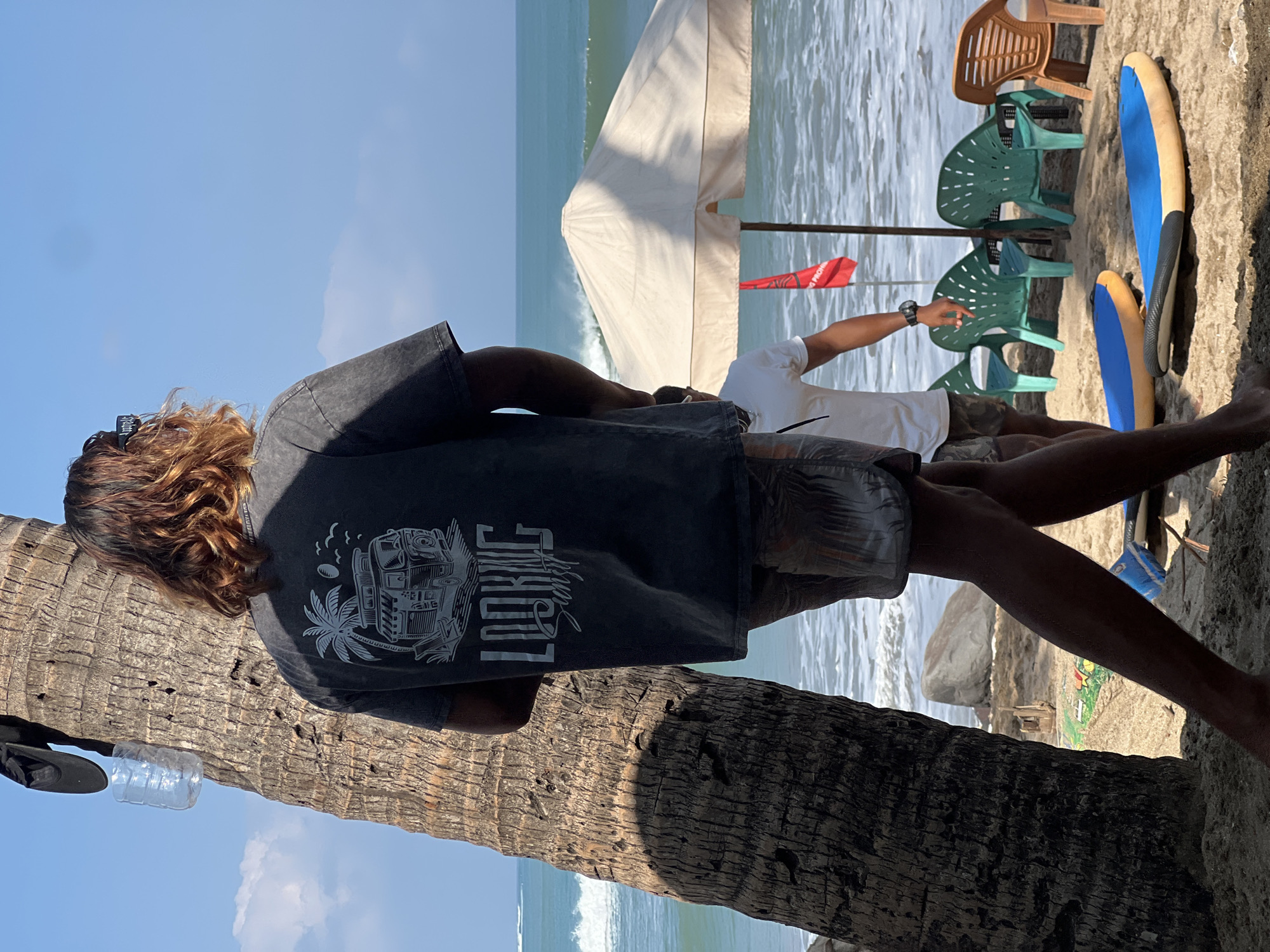 The image depicts a beach scene in Kuta, Indonesia. In the foreground, a person with long, wavy brown hair, wearing a dark grey t-shirt with a graphic print and patterned shorts, stands with their back to the camera, leaning against a large palm tree. A plastic water bottle and a black cap are hanging from the tree.
Further in the background, the ocean with waves is visible under a clear blue sky with some scattered clouds. Several plastic chairs, some green and some orange, are arranged on the sand, under a large, cream-colored umbrella. A red flag with text is planted near the umbrella. Two surfboards, one blue and yellow, and another blue, are lying on the sand in front of the chairs. Another person, wearing a white t-shirt and shorts, is standing near the umbrella, facing away from the camera. The beach is sandy, with some rocks visible near the waterline in the distance.
