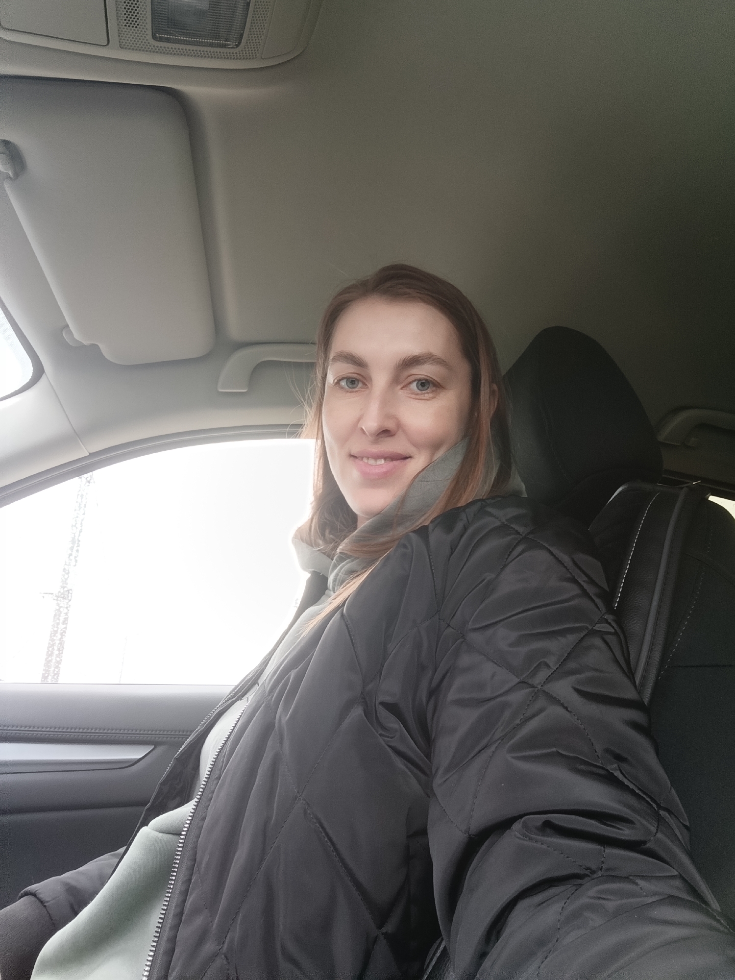 A woman is sitting in the driver's seat of a car. She is wearing a grey hooded sweatshirt and a black quilted jacket. The car is parked, as can be deduced from the partially frosted window to the left of the frame. The sun visor is in the closed position. The setting might be Chaikovsky, Russia, but there are no explicit identifiers to confirm this.