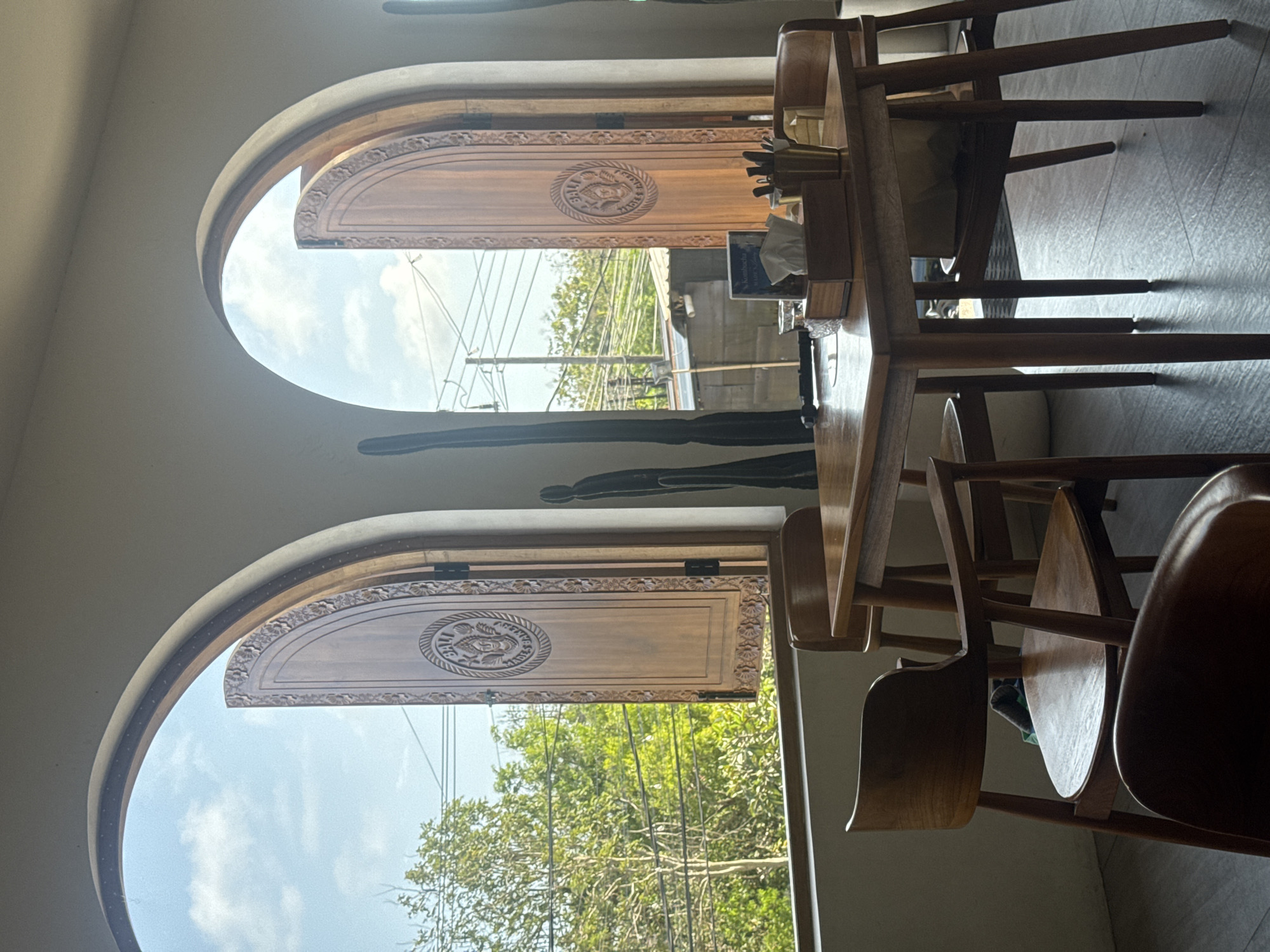 The image depicts the interior of a cafe with two large, arched wooden windows that are open, revealing a bright sky with scattered clouds and lush green trees outside. The windows feature intricately carved wooden shutters with circular emblems. In the foreground, a square wooden table is set with chairs. On the table, there is a wooden tissue box, a glass of water, a small book or menu, and a brass container filled with utensils. Several wooden chairs are arranged around the table. The floor is dark gray. To the right of the right window, a tall, dark decorative object resembling a cactus stands. The scene suggests a serene and inviting cafe environment in Kuta Selatan, Indonesia.