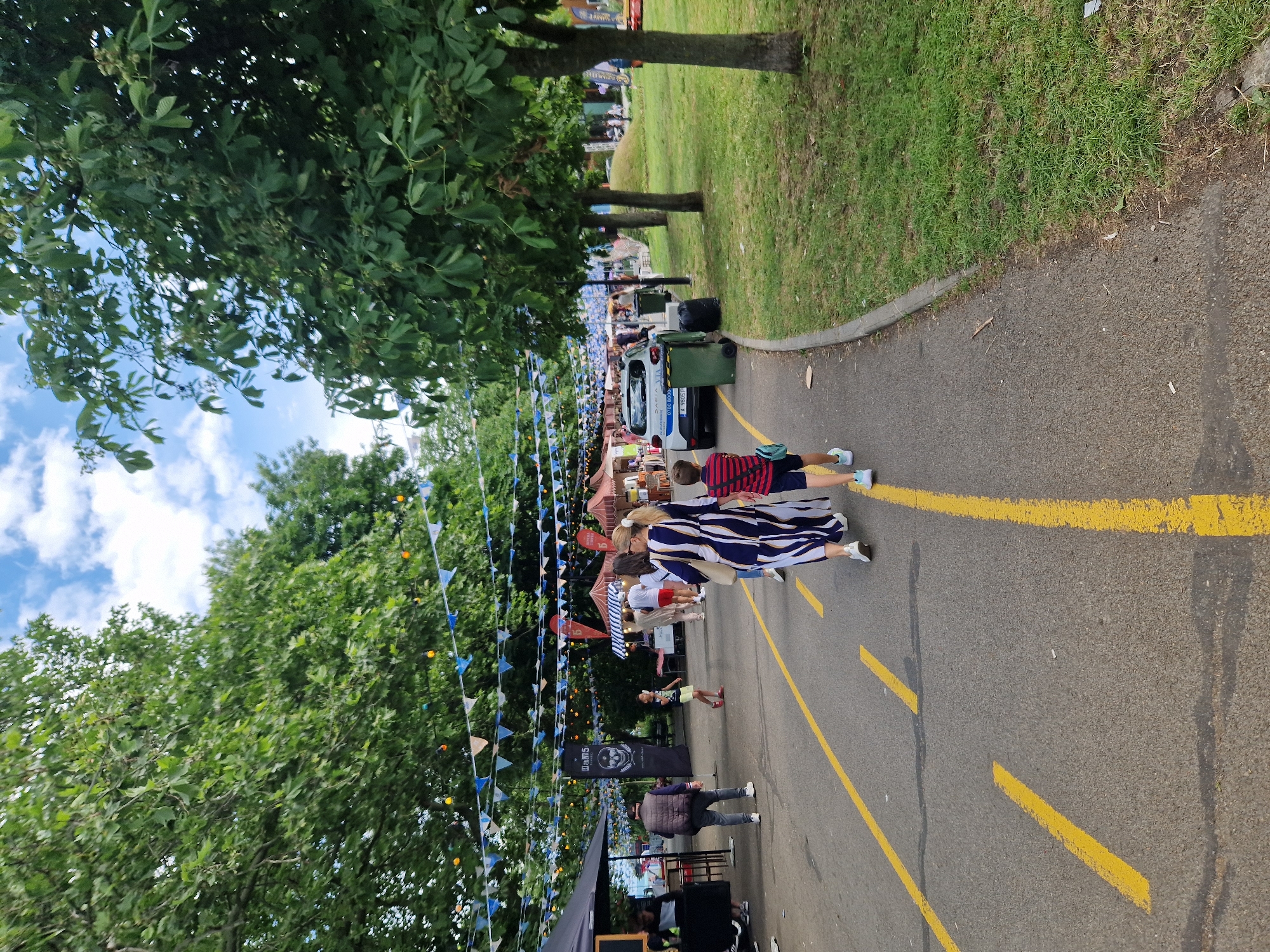 This image captures an outdoor event or market set up on an asphalt road in an unknown city and country, under a partly cloudy sky. The road features yellow dashed and solid lane markings and is flanked by a grey concrete curb on the right, bordering a grassy area with mature deciduous trees. Similar trees are visible on the left side of the road, and decorative blue, white, and light brown bunting, along with strings of small colored lights (orange, blue, yellow), are strung overhead between the trees. On the left side of the road, several vendor stalls or tents are set up. The nearest visible stall is a black tent with a dark tabletop. Adjacent to it stands a tall black banner displaying a white skull graphic and the text "ИДЕАЛ" (or "НАМ5") above additional smaller text. Further along the road, other market tents, some with striped patterns (red and white, blue and white), are visible. Several individuals are present, walking along the road. In the foreground on the right, a boy, estimated to be between 6 and 10 years old, walks away from the camera. He wears a short-sleeved shirt with red and dark blue horizontal stripes, dark blue shorts, and light blue sneakers, and carries a light blue shoulder bag. Slightly ahead and to his left, a woman walks in the same direction. She has blonde hair tied back with a white scrunchie and wears a long, loose-fitting dress with vertical dark blue and white stripes, and white sneakers. She carries a light-colored handbag on her left shoulder. Further down the road on the left, a man is seen from behind, wearing a dark vest over a lighter long-sleeved shirt and dark trousers, also walking away from the camera. A child in a light yellow t-shirt and dark shorts is visible standing or walking in the mid-ground. Numerous other people, adults and children, are distributed throughout the scene, walking among the market stalls. On the right side of the road, a white Dacia Duster vehicle is parked, facing away from the camera. Its rear license plate, "A 9805 P", is visible, along with text "Dacia VIA" and a phone number "0700 8000-" on its rear bumper. Two green wheeled refuse bins and a black trash bag are positioned on the curb next to the parked car. The grassy area on the right is green and well-maintained, with