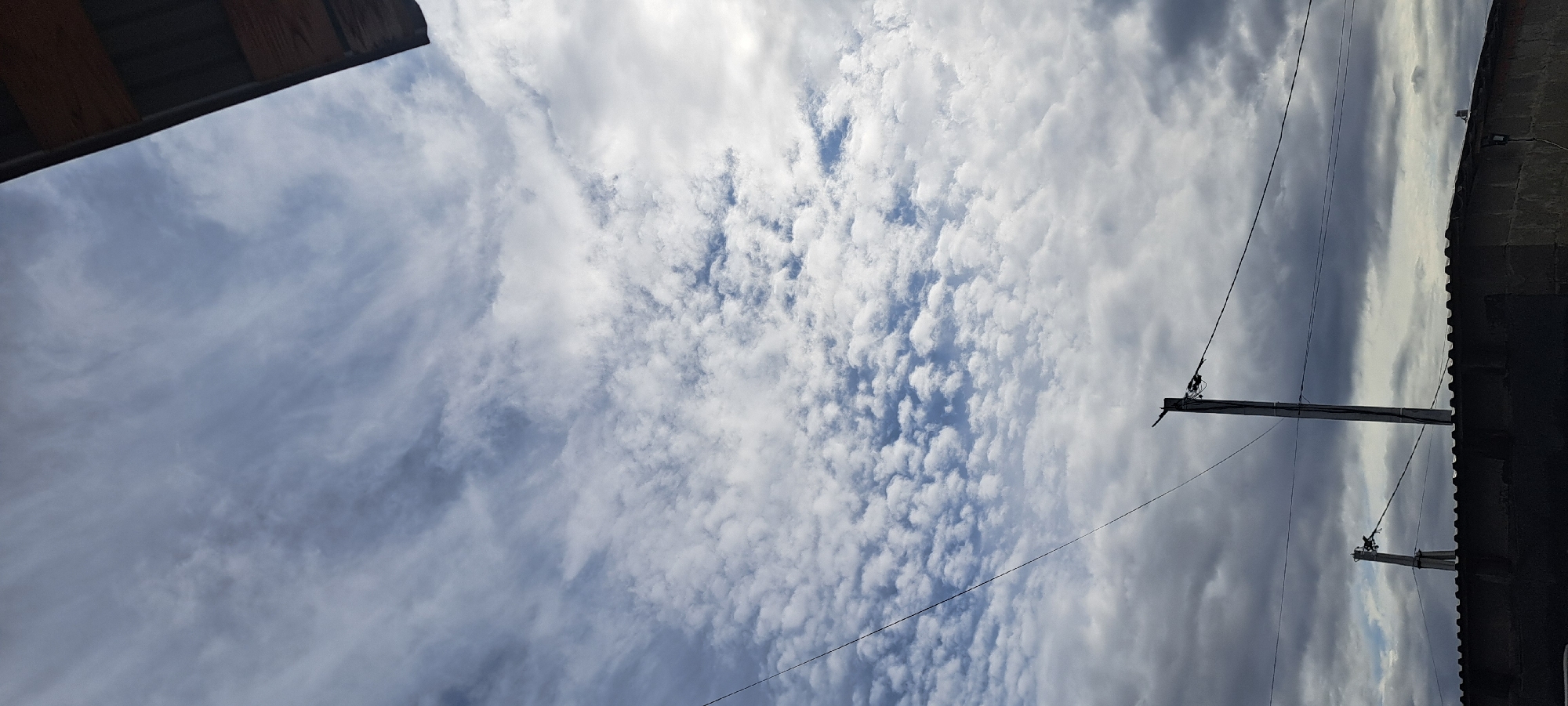 A vertical, outdoor, daytime, low-angle shot captures a cloudy sky above a residential area. The sky is filled with a mixture of thin, wispy cirrus clouds stretching horizontally across the upper frame, and smaller, puffy cumulus clouds in the middle and lower sections. A sliver of a wooden roof structure is visible in the top right corner. A black wire stretches diagonally from the upper left to the middle of the frame. Below the sky, two utility poles with wires are prominent. The pole on the right is taller and more centered in the frame, with several wires attached and a bird visible on its top. The pole on the left is shorter and further back. A dark, low-lying structure, possibly a building or wall with a corrugated roof, runs across the bottom of the image. The scene is set in Azmutkino, Russia.