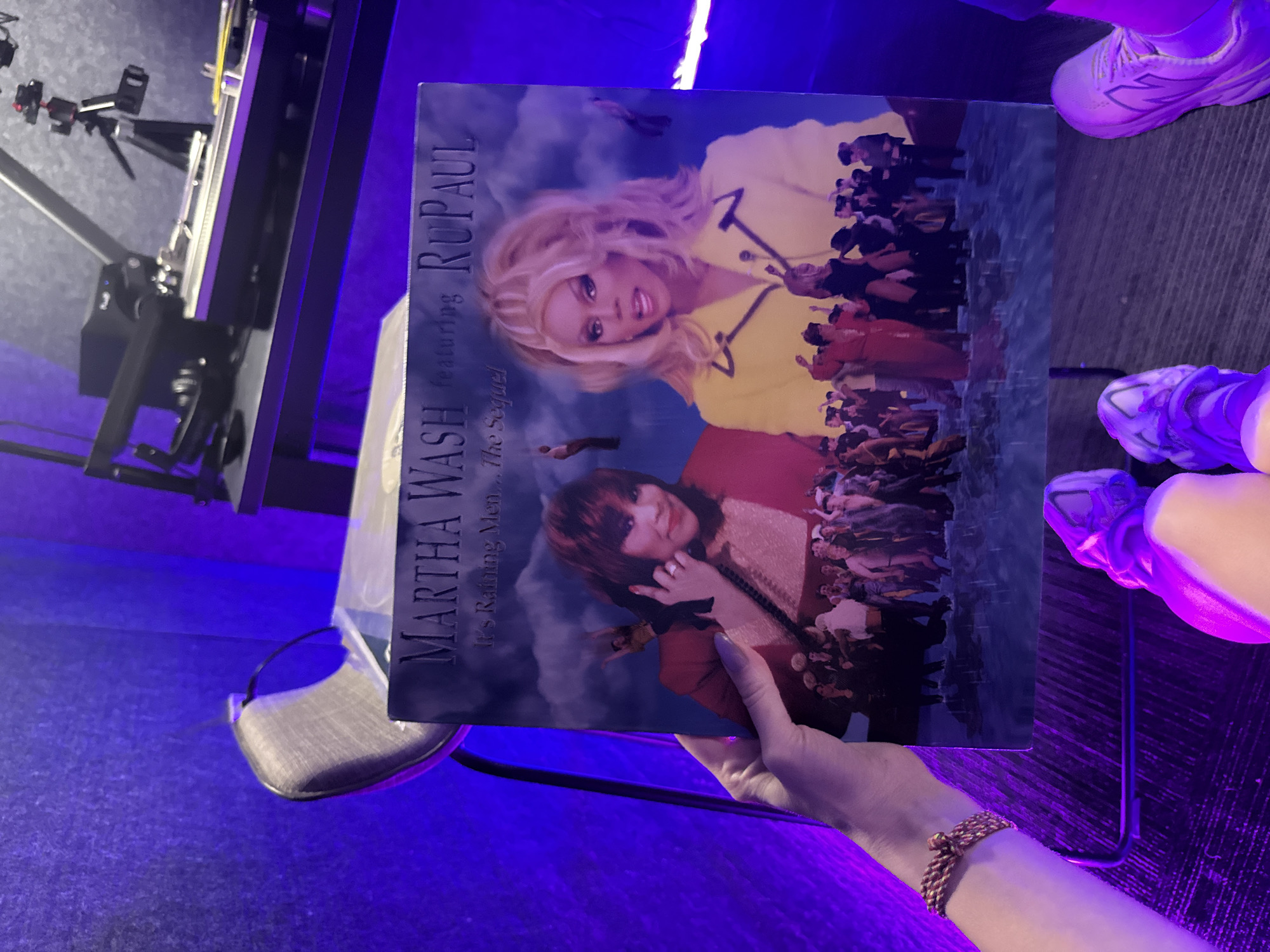 A hand holds a vinyl record album in a room illuminated by purple lighting. The album is "Martha Wash Featuring RuPaul, It's Raining Men...The Sequel". The cover features images of Martha Wash and RuPaul against a sky backdrop filled with figures. A black metal stand with a gray padded seat is visible behind the hand holding the album. A piece of video equipment with a tripod stands to the left of the chair. A person's feet in sneakers are in the upper right corner of the image. The exact location in Jing'an District, China cannot be determined from the provided image.