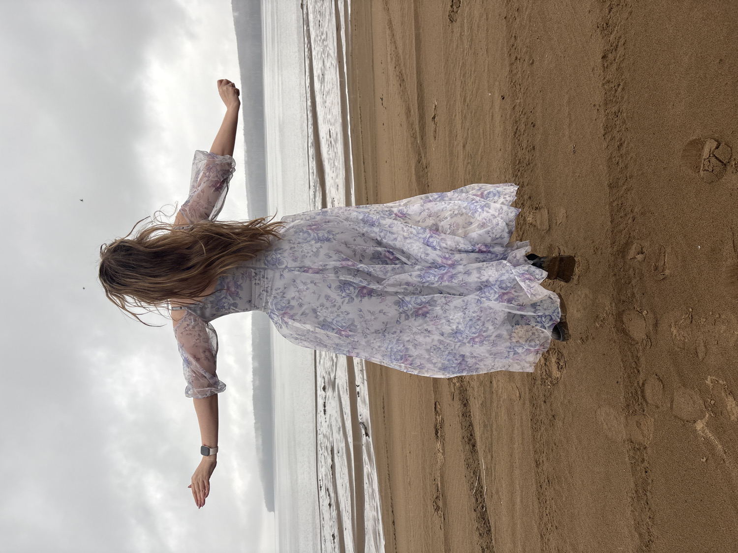 An outdoor, full-length, back view photograph captures a person, presumably a woman, standing on a sandy beach. She faces away from the camera, looking towards a vast body of water under an overcast sky. Her long, brown hair cascades down her back, partially obscuring her head and shoulders.
She wears a light-colored, possibly white or pale gray, dress adorned with delicate purple and blue floral patterns. The dress features sheer, slightly puffed sleeves with cuffed wrists. A smartwatch is visible on her left wrist. Her arms are outstretched to her sides, almost parallel to the ground, suggesting a pose of openness, freedom, or contemplation. Her footwear, possibly dark boots or shoes, is partially visible at the bottom of the dress, covered by the fabric.
The setting is a wide, sandy beach that stretches from the foreground into the midground. The sand shows various textures and shades of brown, with some footprints and small debris visible. To the left, a calm body of water extends towards the horizon, reflecting the muted light of the sky. The horizon line is low, emphasizing the expanse of the water and the large, cloudy, grey sky above. Two small bird-like shapes are faintly visible in the sky to the upper left.
The overall atmosphere is serene and somewhat muted due to the cloudy weather. The location is contextualized as Chaikovsky, Russia, implying the body of water is likely the Kama River or a reservoir. There is no visible text in the image.