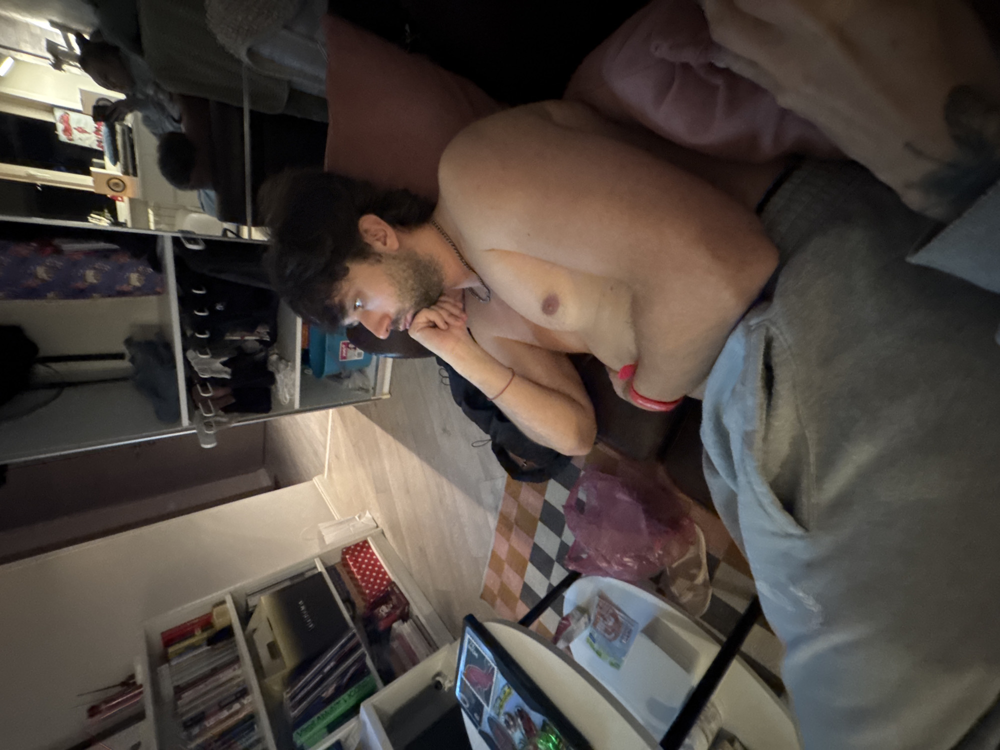 The image depicts a young man sitting on a couch, looking to his left. He is shirtless and wearing grey sweatpants. In the background, a white bookshelf is filled with books and various items. To the right of the man, there is a large mirror reflecting another person and a shelf with clothing. A pink plastic bag rests on the floor near the couch. The scene appears to be indoors, likely in a residential setting. There is no indication of the location being Moscow, Russia.