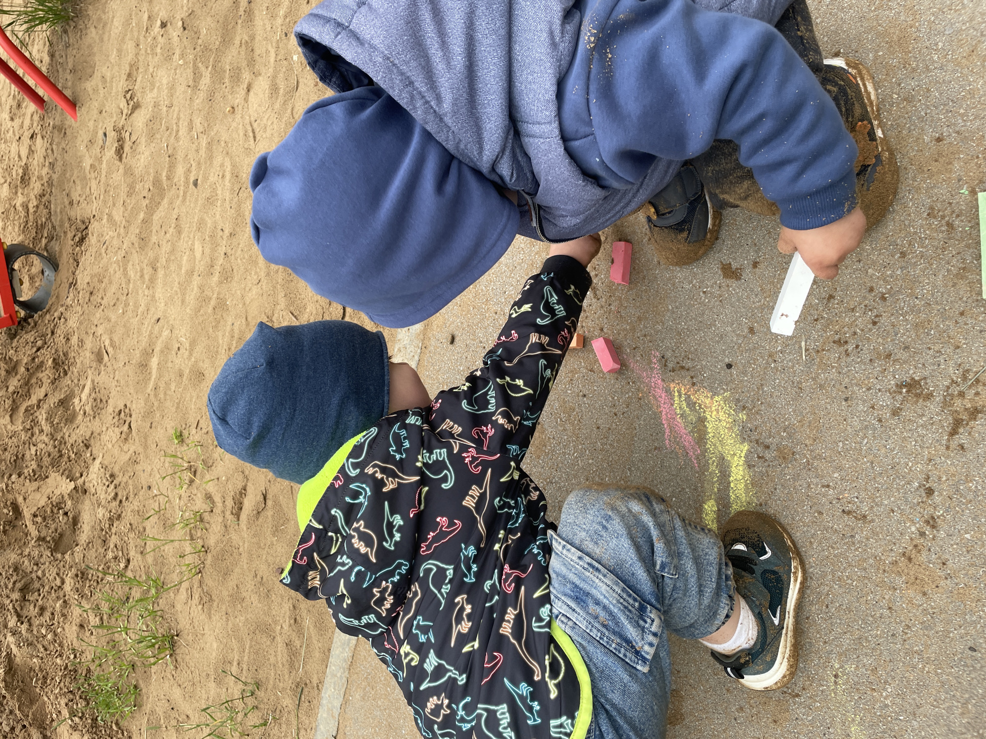The image depicts two young children, likely toddlers or preschoolers, engaged in outdoor play on a concrete and sandy surface in Perm, Russia. Both children are in a crouched position. The child on the left is wearing a black jacket featuring neon-colored outlines of dinosaurs, light blue denim jeans, and dark sneakers heavily soiled with sand. A blue beanie covers their head. Their left arm is extended towards several pieces of chalk positioned on the ground. The child on the right is dressed in a blue hooded jacket with the hood up, blue sweatpants, and dark sneakers also covered in sand. This child holds a white stick of chalk in their right hand, with their left arm also reaching towards additional chalk pieces on the ground. The ground surface shows areas of concrete interspersed with sand and dirt, upon which yellow and pink chalk marks are visible. Two distinct pink rectangular chalk pieces are also present. The background features a large mound of light brown sand, from which sparse green grass shoots emerge. Partial views of red metal playground equipment and a metal wheel-like object are visible further within the sandy area.