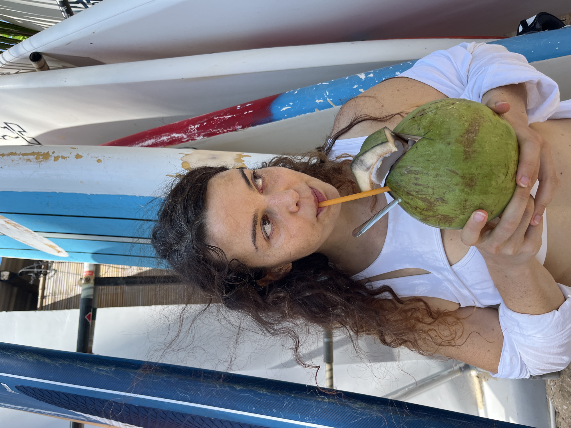 In Phan Thiết, Vietnam, a woman with curly, dark brown hair and fair skin is pictured outdoors, holding a fresh, green coconut with a straw. She is wearing a white top with cutouts and white sleeves. She is looking to her right, away from the camera, and appears to be drinking from the coconut. The background features a collection of surfboards leaning against a structure, with some of them displaying blue, white, and red colors. The environment suggests a beach or a water sports location.