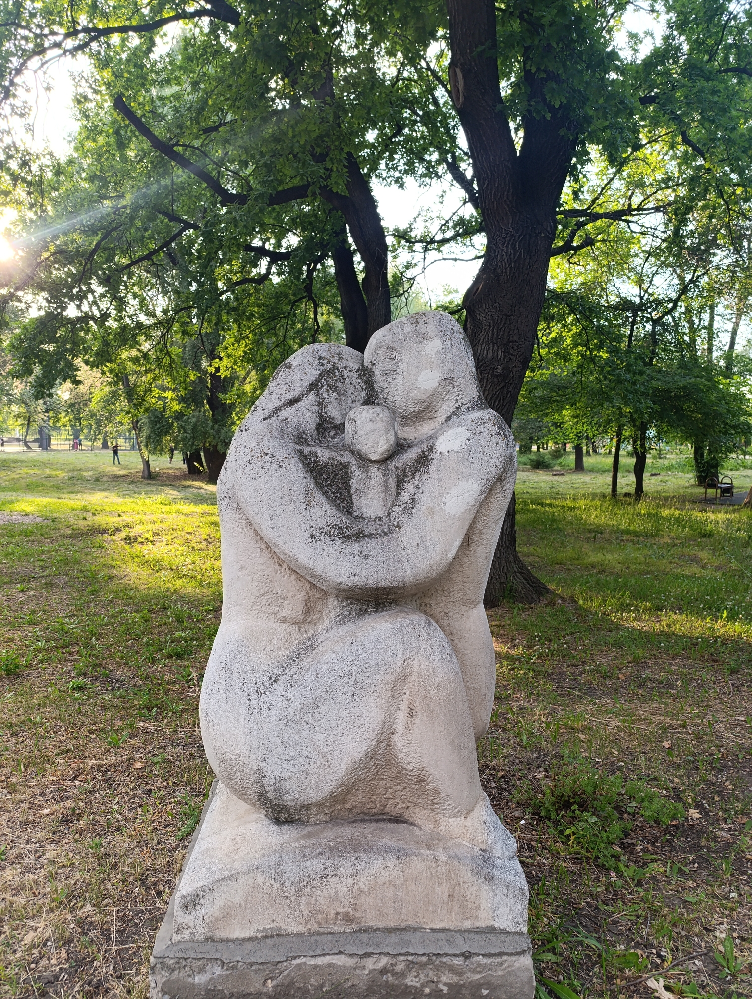 This image depicts a stone sculpture of a family, likely a mother, father, and child, embracing. The sculpture is positioned outdoors in a park setting. The background shows a large tree with dappled sunlight filtering through its leaves, a grassy area with scattered dry leaves and patches of green grass, and other trees in the distance. The sun is shining from the upper left, casting light and shadows across the scene. There are also a few indistinct figures of people in the far background, suggesting a public space.
The specific location of this sculpture within Novokuybyshevsk, Russia, is at the monument to the "Maternity" located in the Memorial Park of the Great Patriotic War. This park is a significant landmark in the city, commemorating the sacrifices of the war.