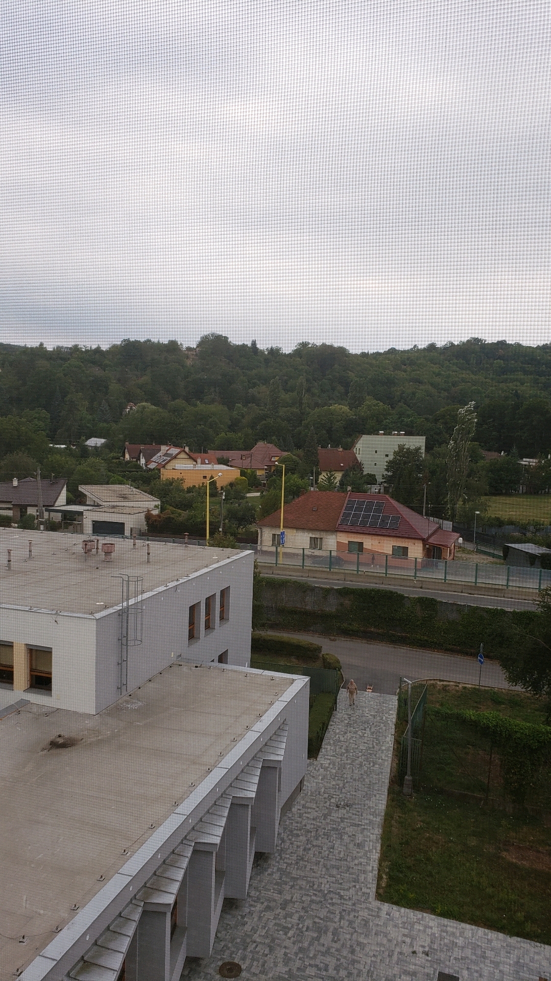 The image shows an overcast day in a residential area with a hilly, forested background. In the foreground, there is a modern building with a flat roof, featuring a prominent grey facade and a series of protruding architectural elements. Adjacent to this building is a paved walkway leading away from it, with a person walking on it. Beyond the walkway, there is a road with a fence and a grassy slope. Further in the distance, several houses with red tile roofs and one with solar panels are visible amongst trees. The overall impression is of a suburban environment with a mix of modern and older residential architecture. This scene is located in the city of Prešov, Slovakia, specifically in the vicinity of the residential area called Sekčov.