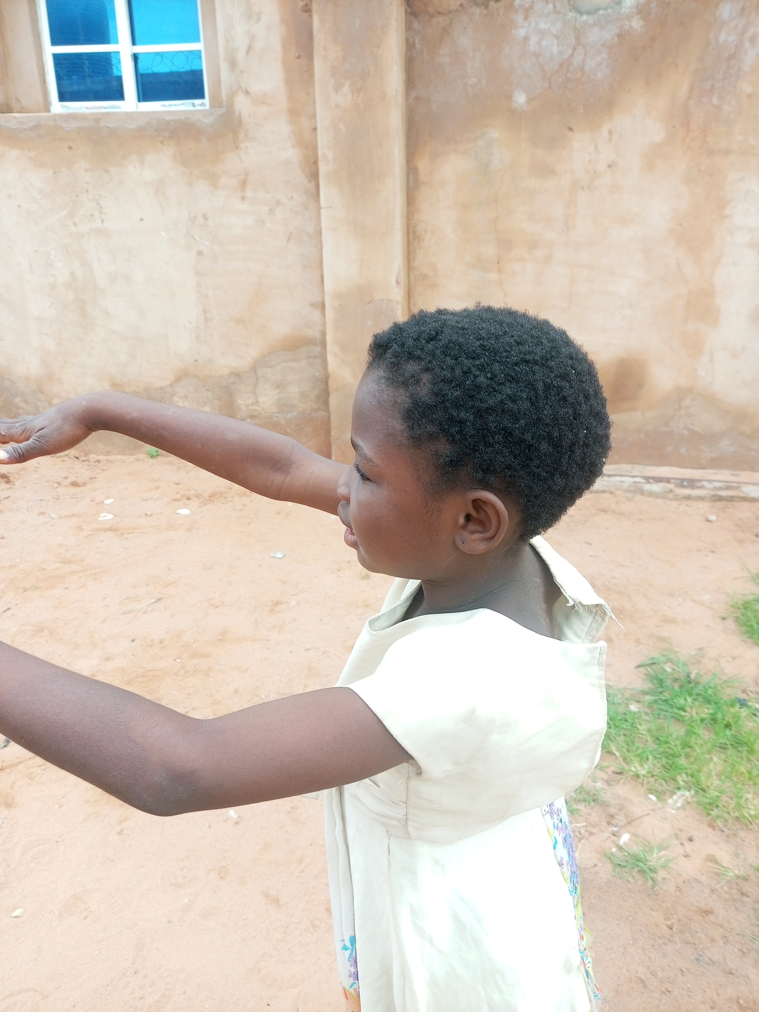 A medium shot captures a young Black girl in an outdoor setting, likely in Tempure, Nigeria. She is wearing a light-colored dress with floral accents and has short, curly black hair. Her head is turned to her right, with her face visible in profile, and her mouth is slightly open. Her left arm is extended forward and slightly upward, with her hand open. The background consists of a sandy ground, a textured, tan-colored wall with some discoloration, and a window with blue panes. The environment suggests a casual, possibly residential area.