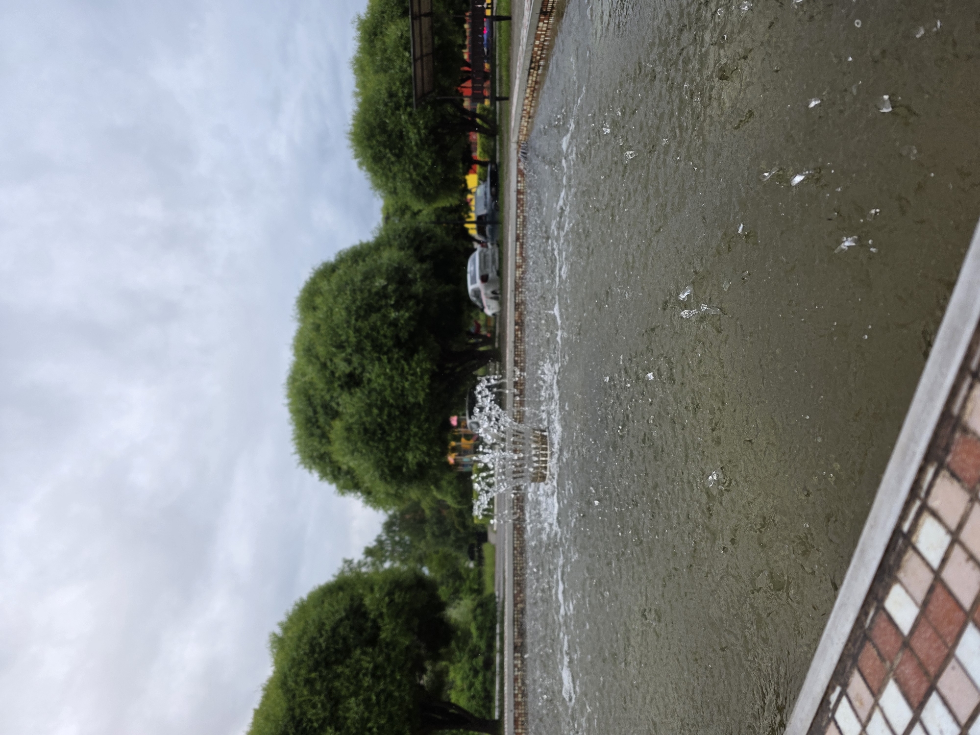 This vertical image depicts a public park setting, stated to be in the city of Chaikovsky, Russia, on an overcast day. In the foreground, the edge of a large, rectangular fountain basin is visible, featuring a tiled border composed of small square tiles in shades of brown, white, and reddish-brown, capped by a light gray coping. The water within the fountain is a murky greenish-brown, with numerous water droplets actively splashing and falling back into the basin. A single, distinct water jet erupts from a brass-colored nozzle located in the mid-left portion of the fountain, forming a concentrated, crown-like splash. Beyond the fountain, a paved area or road stretches horizontally, bordered by a row of evenly spaced, deciduous trees characterized by dense, round green canopies. In the background, several vehicles, including a white sedan and a darker car, are visible on the paved surface. Further back and to the right, indistinct colorful structures, possibly associated with a playground or recreational facility, are present, alongside a covered shelter. The sky above is uniformly overcast with light gray clouds, providing soft, diffused illumination to the entire scene. There are no individuals explicitly visible in the frame.