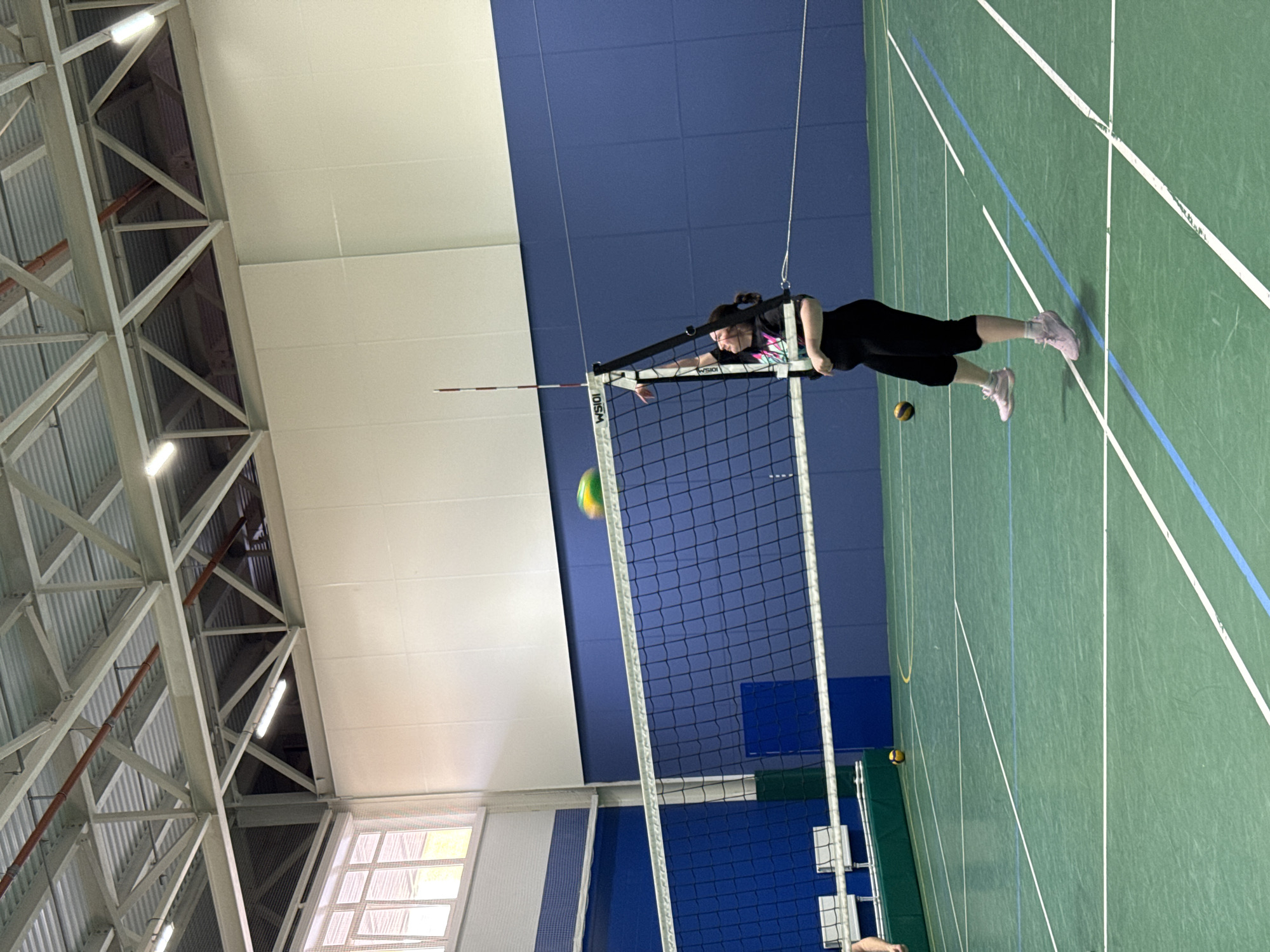 A young woman is playing volleyball indoors. She is in mid-air, jumping to hit the ball, which is blurry as it is in motion. She is wearing black leggings, a black t-shirt with a colorful graphic, and pink sneakers. The volleyball net is in the foreground, and a volleyball is also visible on the green court in the background. The walls are painted blue and white, and the ceiling has exposed beams and lighting. The image appears to have been taken at a sports facility in Saint Petersburg, Russia.