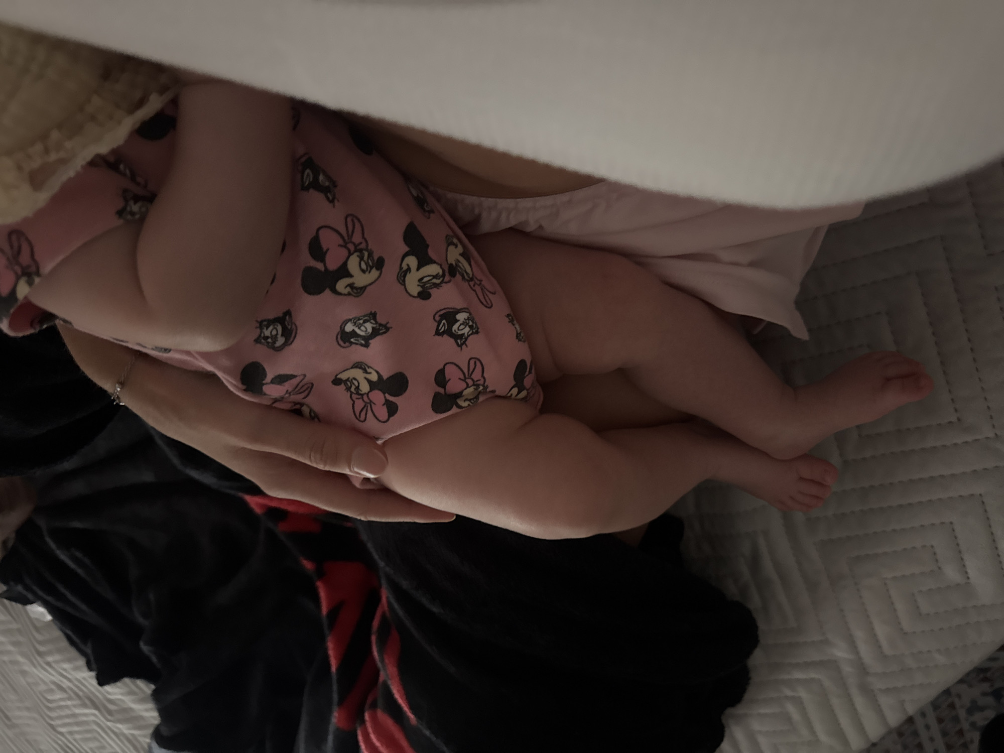 The image shows a close-up view of a baby's legs and feet, resting on a quilted surface. The baby is wearing pink pajamas with a pattern of Minnie Mouse characters. A hand, presumably belonging to an adult, is resting on the baby's leg, and a bracelet is visible on the wrist. The background consists of dark fabric and the quilted surface, suggesting an indoor setting. There is no information in the image that would indicate the location in the city of Isola delle Femmine, Italy.