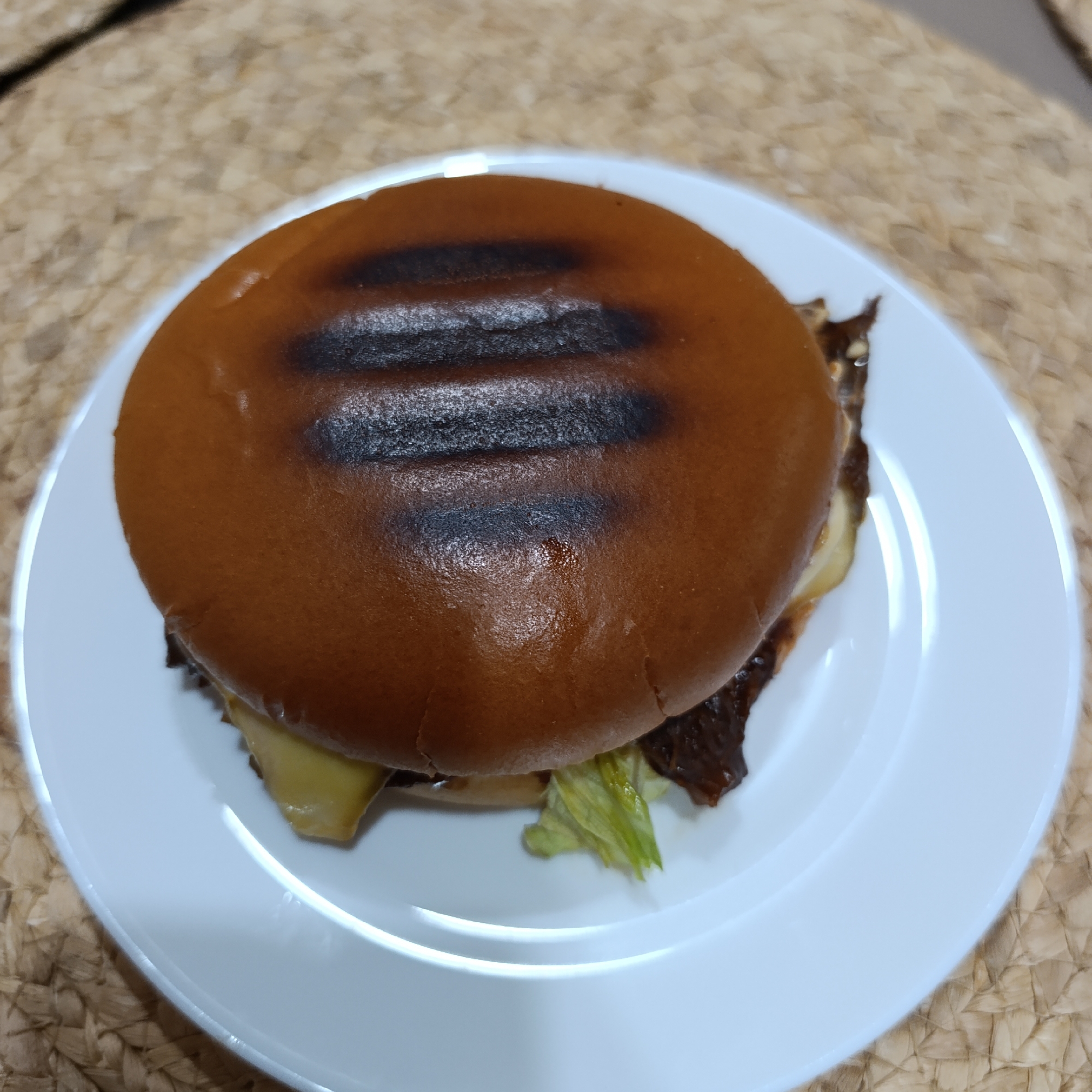 The image depicts a hamburger on a white plate, resting on a woven surface, possibly a placemat or table covering. The hamburger consists of a toasted bun with grill marks, a patty, cheese, lettuce, and possibly other condiments. The location is not discernible from the provided image, and there is no information to suggest it is in the city of Марковский, Russia.