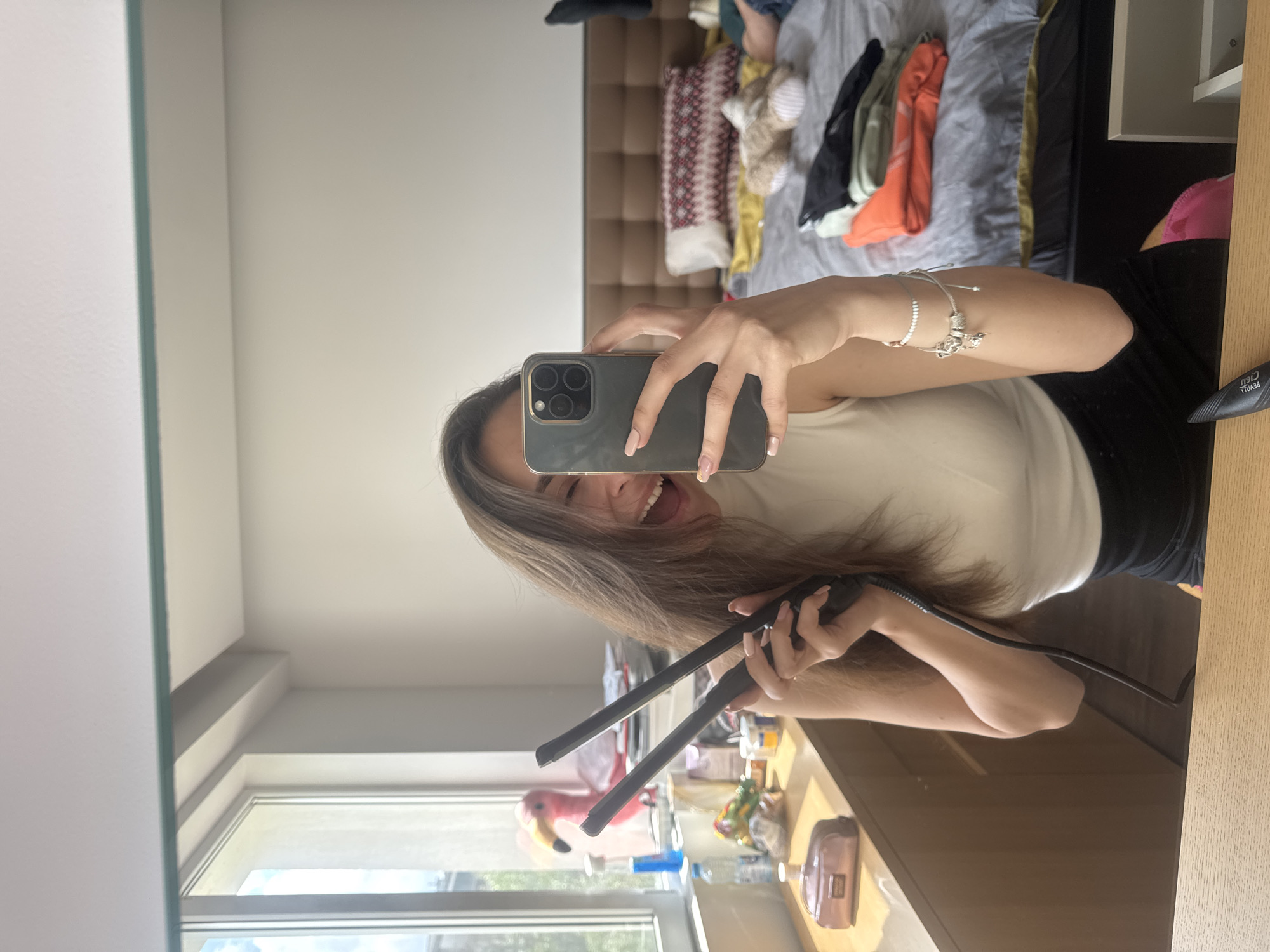 A young woman is taking a selfie in a room. She is holding a black smartphone in her left hand and a black hair straightener in her right hand. She is smiling and looking at the camera. She is wearing a light beige top and black pants. On her left wrist, she is wearing multiple bracelets. The room appears to be a bedroom with a bed in the background. The bed has a headboard and is covered with a grey duvet and several pillows. There is a window in the background with a glimpse of trees outside. On a table near the window, there are several items, including a pink flamingo decoration and a pink purse. The location cannot be determined with certainty to be Sofia, Bulgaria from the visible elements in the image.