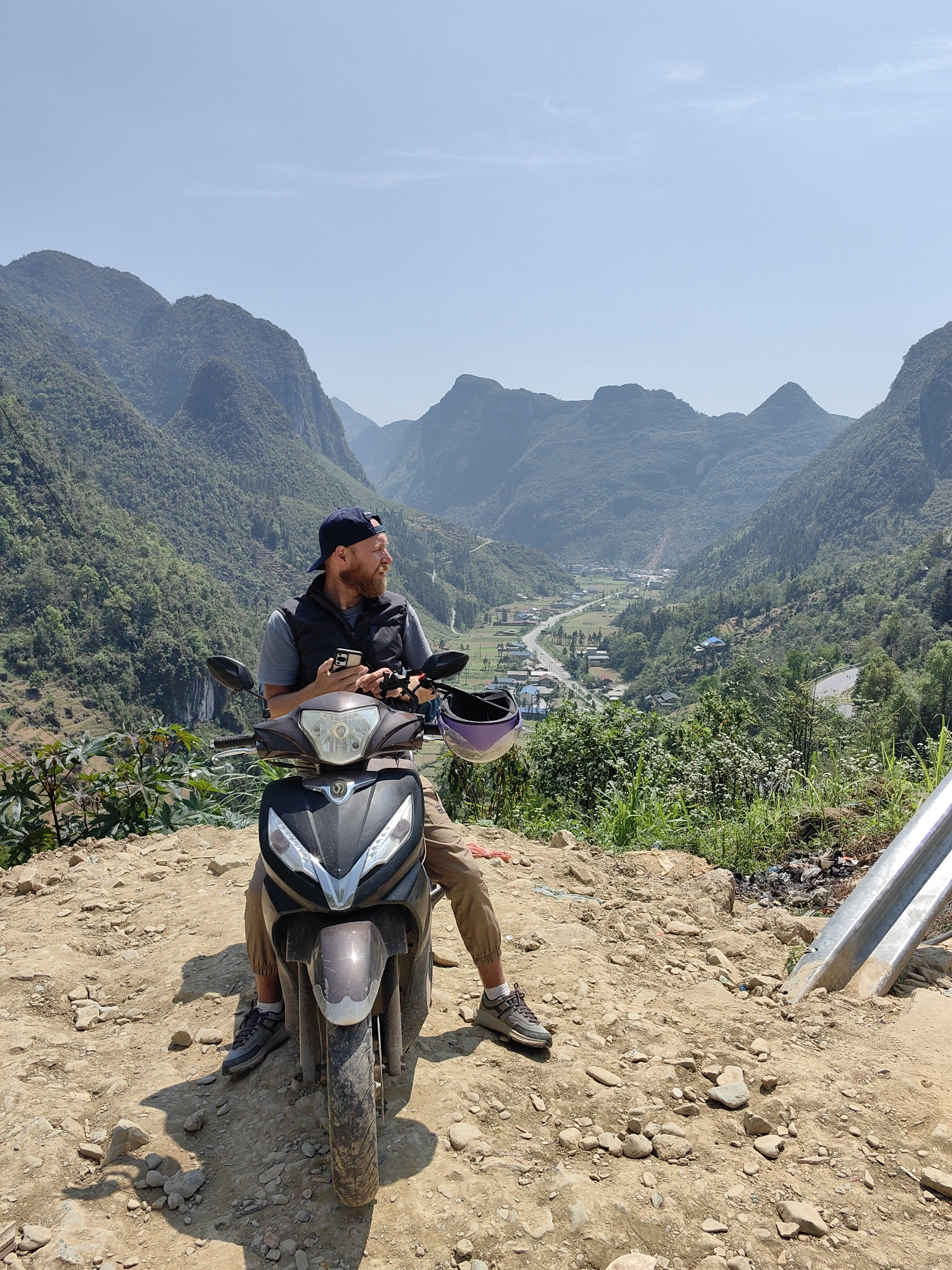 A man sits on a moped on a rocky, dirt-covered incline overlooking a valley, likely in the Phố Bảng Commune, Vietnam, based on the mountainous terrain and the valley community visible in the background.
The man, fair-skinned with a beard and wearing a navy baseball cap, gray t-shirt, and a dark vest, is seated on the moped, looking off to his left. He holds a cell phone in one hand. His legs are khaki pants, and he wears gray sneakers. A purple helmet is seen resting near the moped's handlebars.
The moped is a dark color, mostly black and gray, and is positioned on the dirt path. The foreground is rocky and sparsely vegetated with some small plants. A metal guardrail is visible at the edge of the path on the right side of the image.
The valley beyond features a town or community with buildings and a road winding through it. Mountains covered in green vegetation rise in the background under a clear, blue sky. The overall scene suggests a scenic overlook on a hillside.