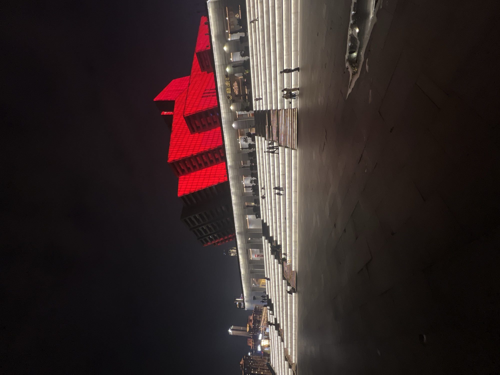 At night, the exterior of a modern building is illuminated by bright red lights on its upper levels, creating a striking contrast against the dark sky. The building features angular, geometric architectural elements. In front of the building, a wide, multi-tiered staircase is brightly lit with white lights along each step, creating a luminous pathway. Several small groups of people are visible on the illuminated stairs and in the plaza area in front of them, appearing to be casually walking or standing. In the background, city lights from other buildings are visible, indicating an urban setting. The ground in the foreground appears to be paved and reflects some of the ambient light. The overall impression is one of a vibrant, modern urban public space at night.