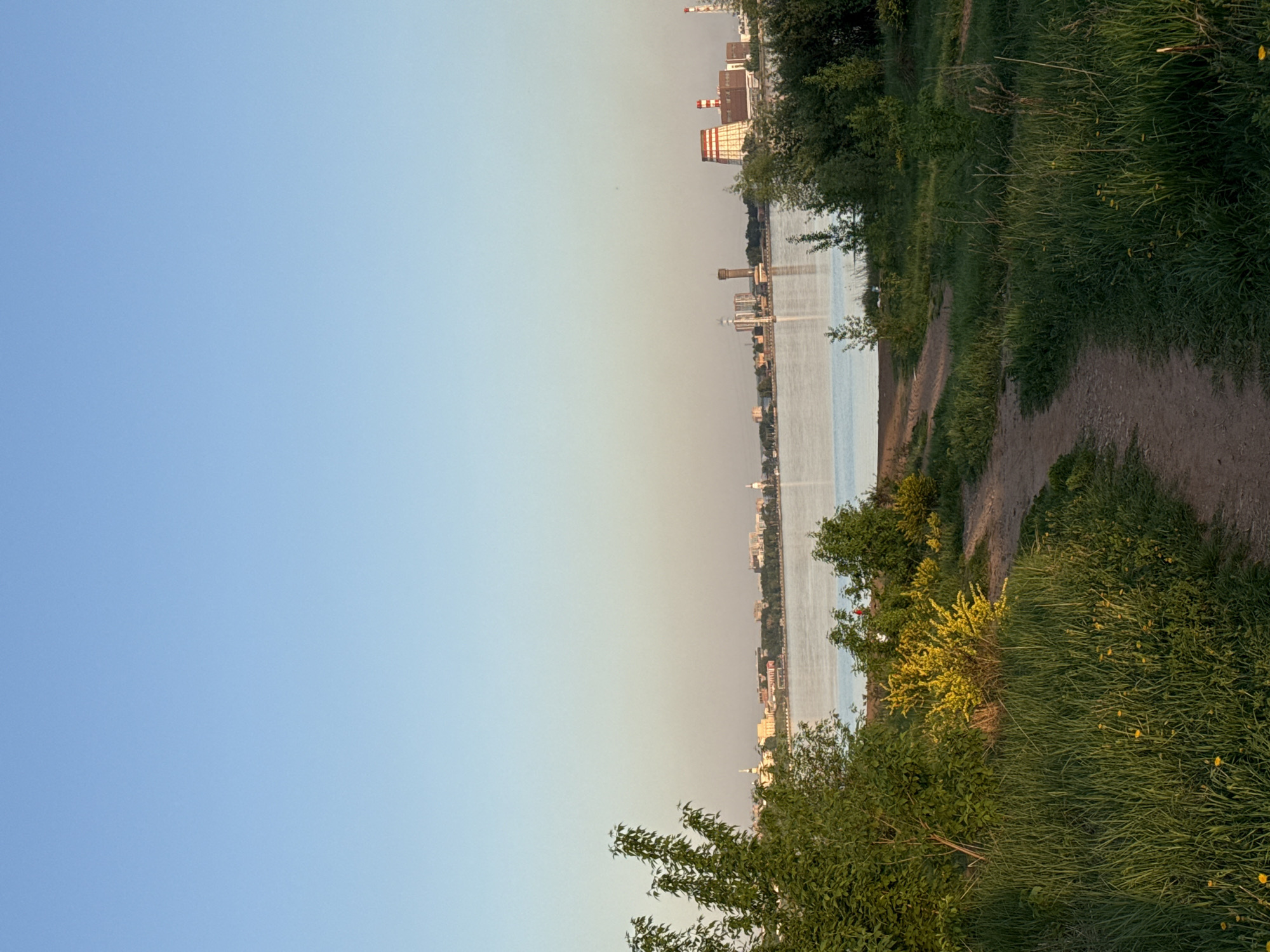 The image shows a landscape in Izhevsk, Russia, likely taken from a park area overlooking the Izhevsk pond. The scene features a path bordering the water's edge with grassy vegetation and bushes along the side. The Izhevsk pond is in the middle ground, showing the calm water reflecting the sky, creating a division between the sky and land. The city skyline is visible on the opposite bank of the pond, marked by industrial buildings and residential areas in the background. The sky is clear and light blue, suggesting either early morning or late afternoon light.