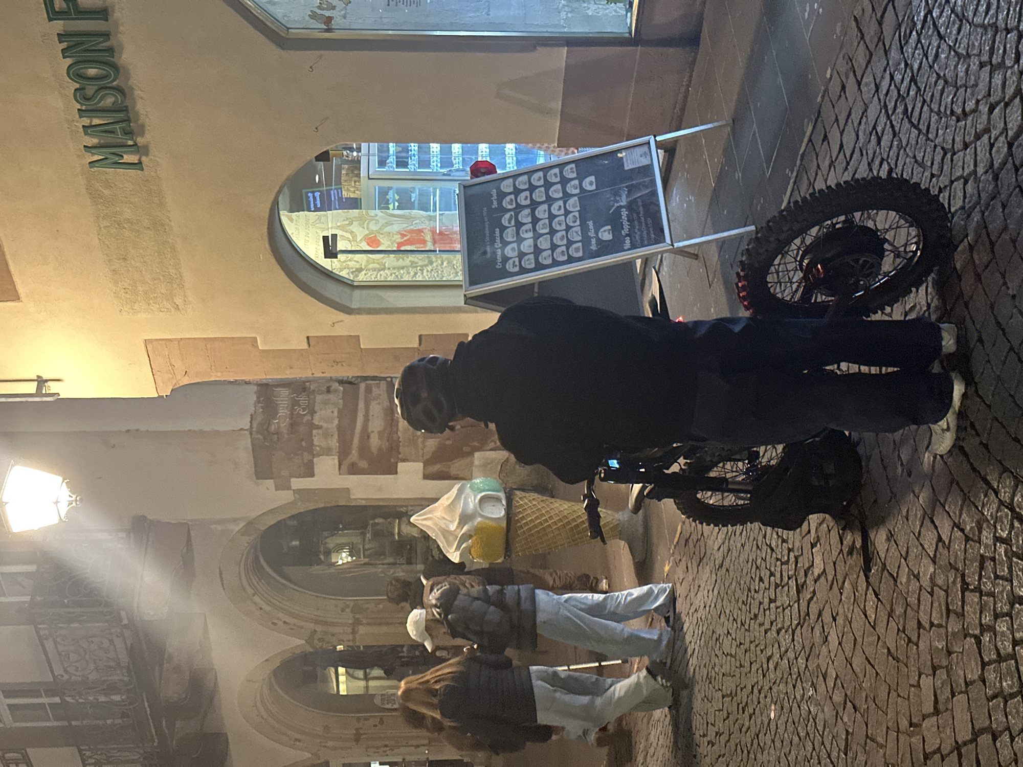 The image captures a street scene at dusk in Strasbourg, France. A person wearing a helmet and dark clothing stands with a motorized bicycle near a storefront. The storefront features a menu board with various ice cream flavors listed, and "MAISON F" is visible on the building above. A small group of people walks past an oversized ice cream cone decoration nearby. The street is paved with cobblestones. A lamppost provides illumination, casting light onto the street and nearby buildings.