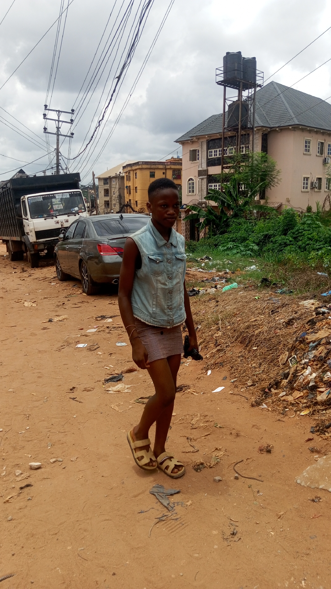 A young person stands on a dirt road in Obosi, Nigeria. The person is wearing a denim vest, a striped skirt, and sandals. A black object is held in one hand. Behind the person are a parked truck, a grey car, and a building with water tanks on the roof. Debris is scattered on the road and near the roadside vegetation. Electrical lines run overhead.