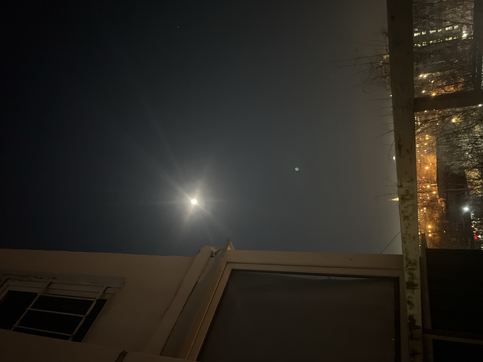 The image shows a night scene, possibly in Saint Petersburg, Russia, although there is no explicit proof in the image. A bright celestial object, likely the moon, dominates the upper portion of the frame. Below, there is a light-colored building facade, with a window visible. To the right, a vertical support structure, possibly a metal beam, is partly visible. In the background, lights suggesting a city skyline are seen through a window or opening in the beam. The overall lighting suggests it is nighttime. The scene appears still and devoid of people.