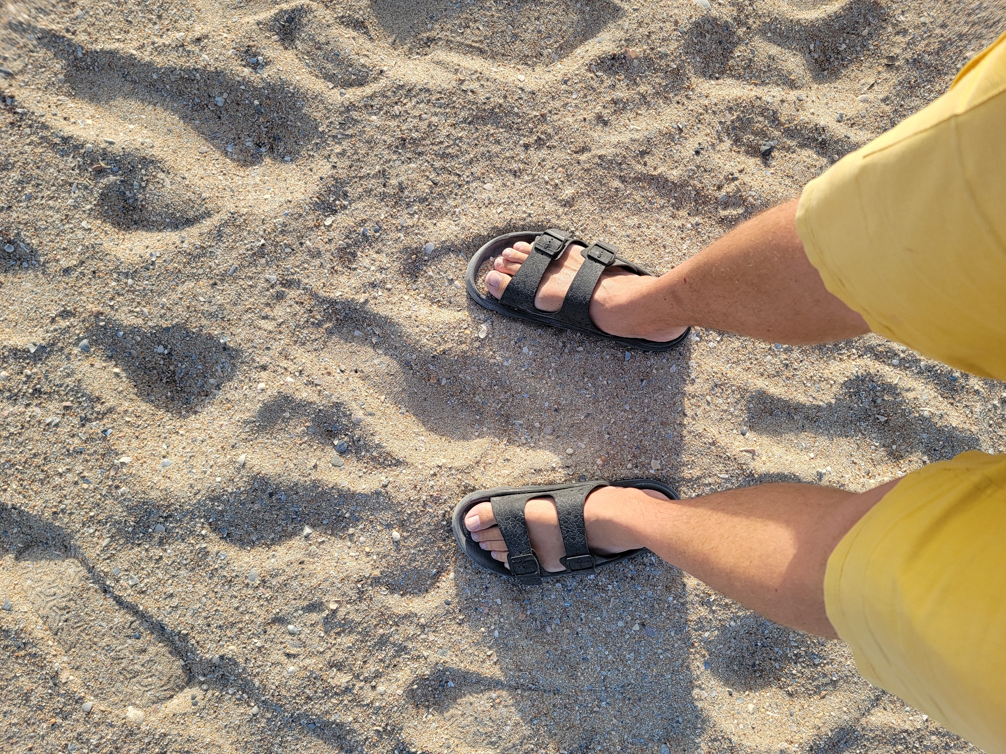 This is a first-person, overhead shot of a person standing on a sandy beach. The person is wearing dark, double-strap sandals and bright yellow shorts. The sand is light brown with small pebbles and shell fragments, and there are shadows cast on it, suggesting sunlight. The background is out of focus, but the texture of the sand is clear. The scene is consistent with a location on a beach in Quarteira, Portugal, known for its sandy shores.