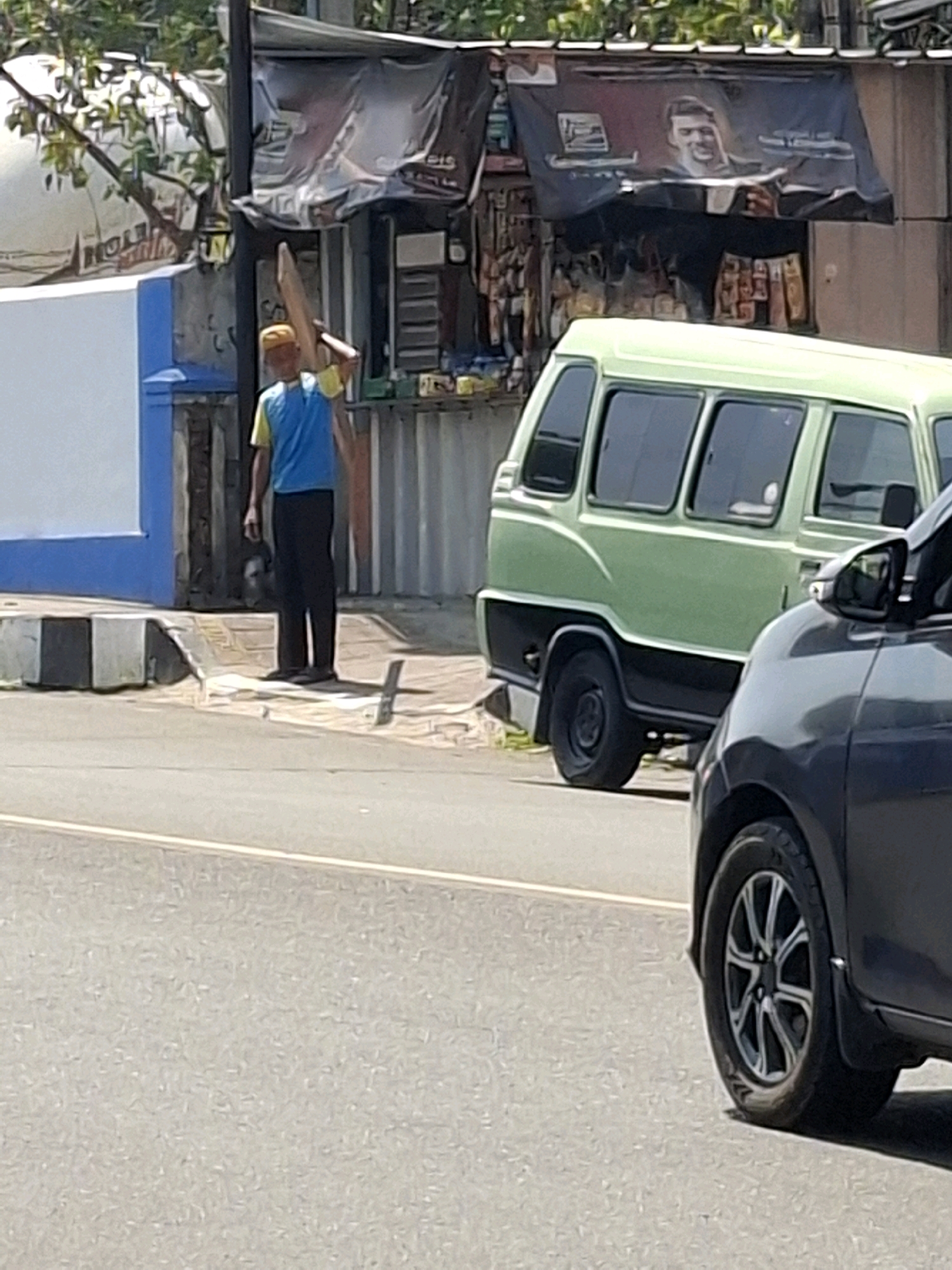 A person wearing a yellow cap, blue and yellow shirt, and black pants stands on a sidewalk, holding a long, light-colored wooden plank over their right shoulder. The person is positioned in front of a small open-air store with shelves stocked with various items. A light green minivan is parked to the right of the store. A dark gray car is partially visible in the foreground, to the right of the minivan. The scene appears to be a street in the city of Cianjur, Indonesia, as indicated by local storefronts and vehicles.