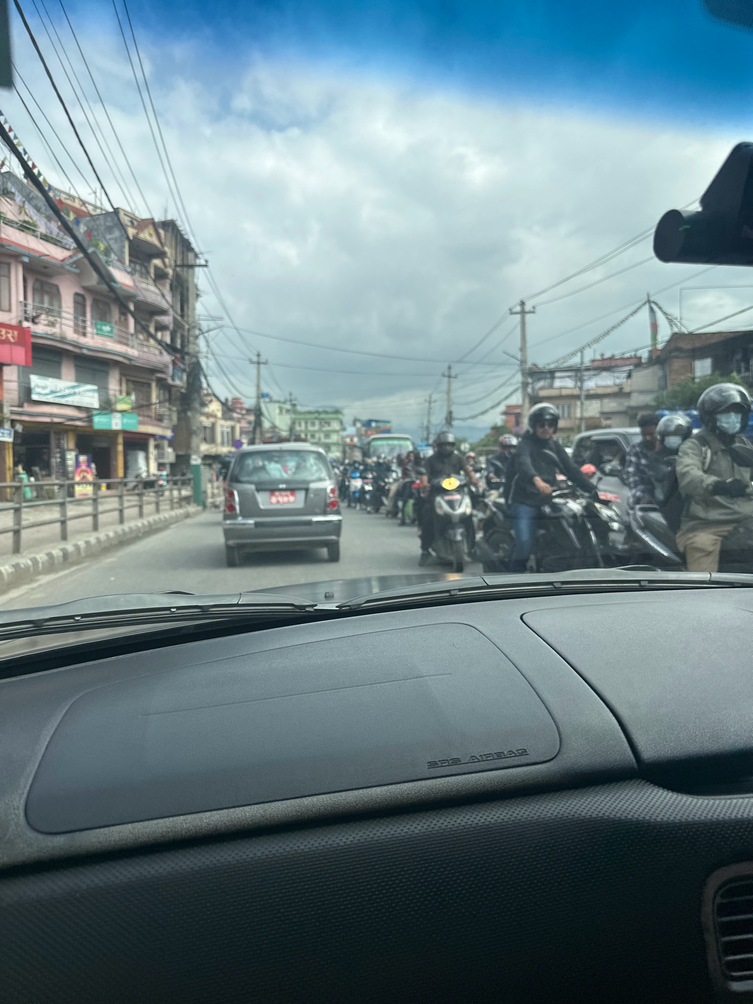 The image depicts a traffic jam on a street in Kathmandu Metropolitan City, Nepal, as viewed from inside a vehicle. The street is lined with multi-story buildings on the left, exhibiting a mix of architectural styles, with signage in Nepali. Numerous motorbikes and a few cars are stationary or moving slowly in the traffic. People on the motorbikes are wearing helmets and casual clothing. The sky is overcast with clouds. Utility poles and wires are visible overhead, common in urban Nepalese environments. The foreground shows the dashboard of the vehicle from which the photo was taken, including the "SRS AIRBAG" indicator.