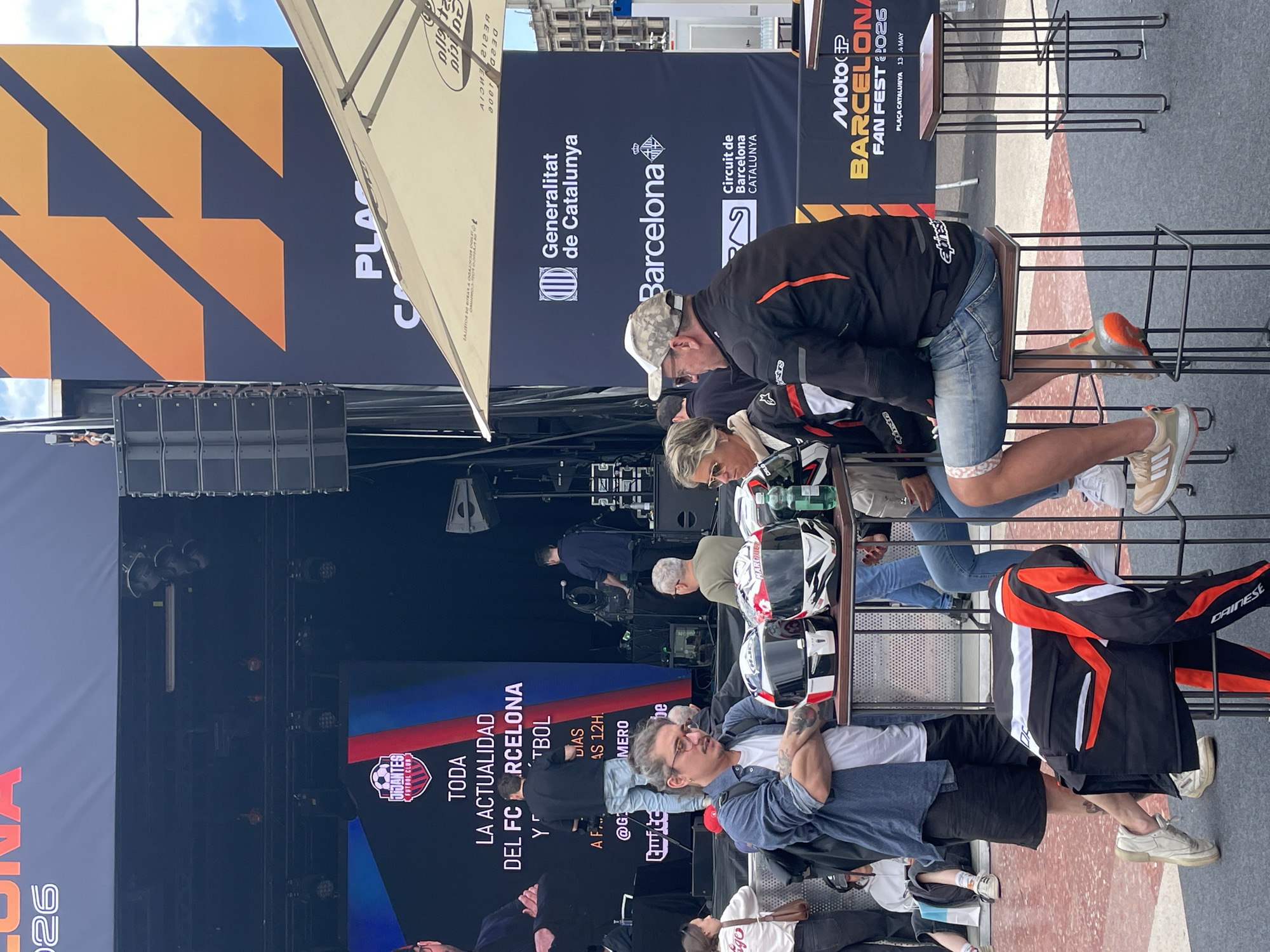 The image shows a scene in Plaça Catalunya, Barcelona, Spain, during the Moto GP Barcelona Fan Fest 2026 event (13th-14th of May). A stage is visible with the "JIJANTES FUTBOL CLUB" logo and a screen displaying information about FC Barcelona. Several people are near the stage, some wearing motorcycle gear and holding helmets. A man wearing a baseball cap and jean shorts is sitting on a metal structure. There is a banner with logos of Generalitat de Catalunya, Barcelona, and Circuit de Barcelona-Catalunya. Other banners are also present, including one with the "PLA" caption and patterns, and another one that reads "Alicia".
