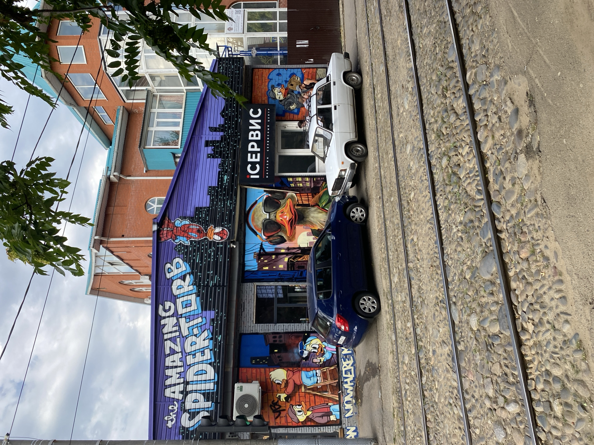 The image shows a street in Krasnodar, Russia, with a building facade decorated with graffiti. The graffiti features "The Amazing Spider-Torrb" lettering and an image of Spider-Man hanging upside down. Other murals include cartoon ducks painting and wearing sunglasses, and a large duck face with sunglasses. A sign above a storefront reads "iСЕРВИС" (iSERVICE) with smaller text below indicating "МАГАЗИН СЕРВИСНЫЙ ЦЕНТР" (SHOP SERVICE CENTER). Two cars are parked on the street: a dark blue hatchback and a white sedan. The street is paved with cobblestones, and tram tracks run across it. Trees with green leaves are visible in the upper right corner, obscuring parts of the building. The sky is cloudy.