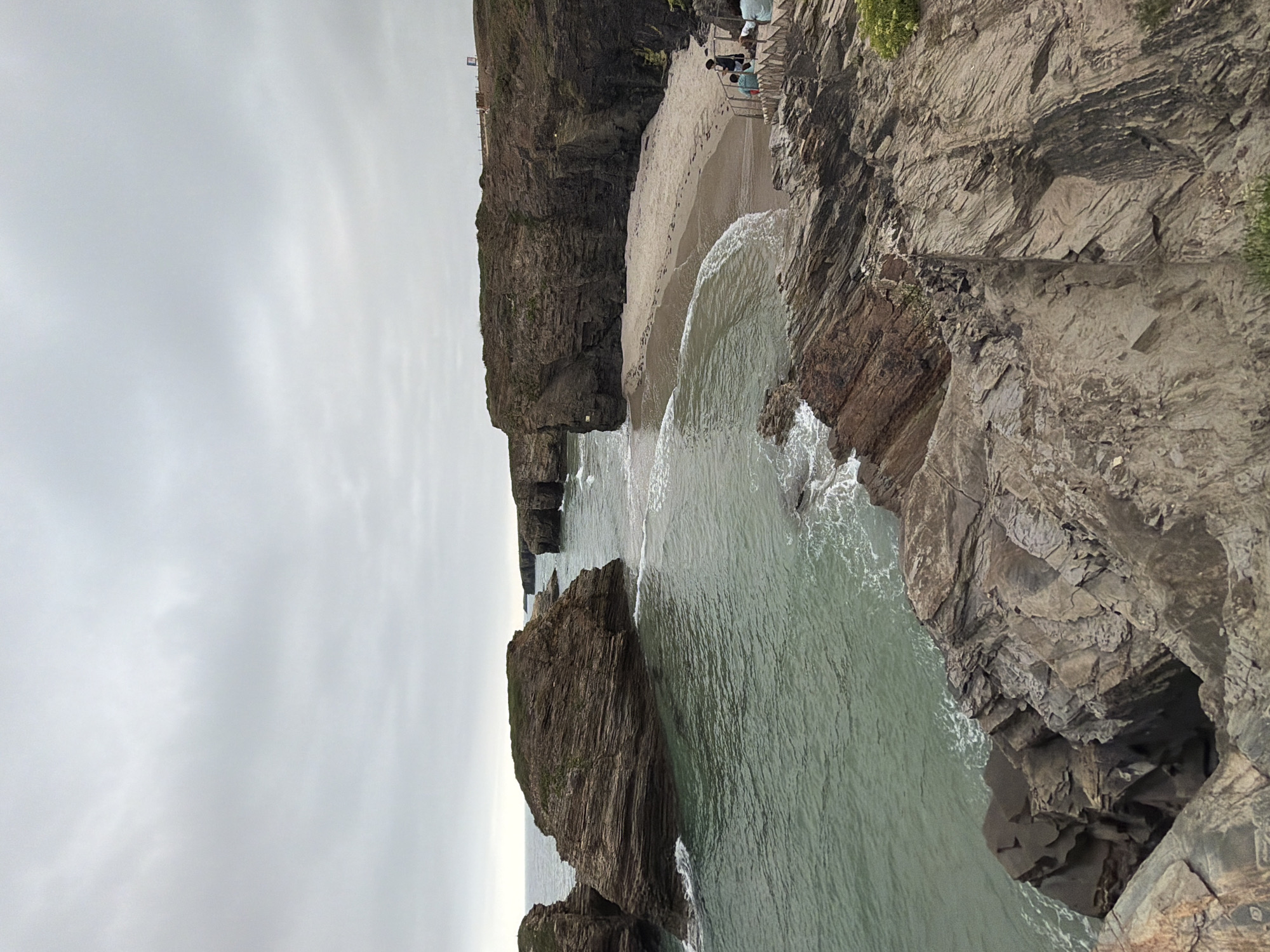 This is a photograph of the Praia de Augas Santas (Beach of the Holy Waters) in Ribadeo, Spain, under an overcast sky. The beach features a crescent of light-colored sand nestled between dramatic, dark, rocky cliffs. Jagged rock formations jut out into the calm, greenish-blue ocean, with small waves lapping at their bases. Footprints are visible on the wet sand near the water's edge.
In the foreground and middle ground, the rocky cliffs are layered and eroded, showing a rough texture. Vegetation, including small green shrubs, grows sparsely on some of the cliff faces. A set of wooden stairs leads down to the beach on the right side of the image.
On the beach, near the stairs, three individuals are present. One person is standing and looking towards the sea, while two others are seated on the stairs, one appearing to be a child. Further down the beach, a small group of people, possibly two or three, are walking. A fence runs along the top of the cliff on the right side of the frame, and a sign is visible on a pole. The overall atmosphere is serene and natural, with the dramatic landscape dominating the scene.