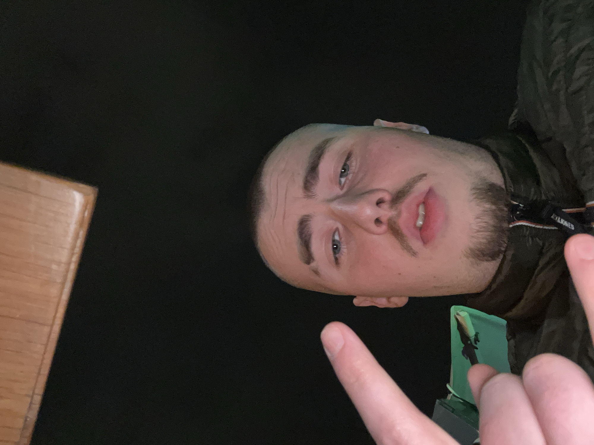The image shows a man with short, light brown hair and a beard looking down at the camera. He is wearing a dark jacket. His index and middle fingers are visible, pointing towards the bottom of the frame. A green and black object is visible near his hand. The background is dark, and a piece of light brown wood is visible in the bottom left corner. The location is unknown.
