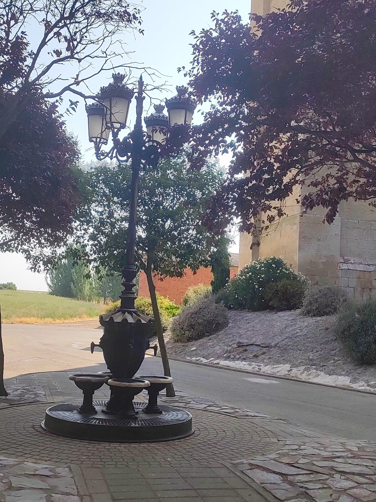 The image depicts a street scene in Autillo De Campos, Spain, featuring an ornate, dark metal fountain with multiple basins in the foreground. The fountain is situated on a paved circular area with intricate brickwork. Behind the fountain, a tall, dark metal lamppost with four lamps crowns the structure. In the background, a paved road runs alongside a slightly elevated, rocky embankment with sparse vegetation. To the right of the embankment, a light-colored stone building, likely a church or historical structure, is partially visible. Lush green trees with deep red leaves frame the upper portion of the scene. The sky is bright and clear, suggesting daytime. No people are present in the image.