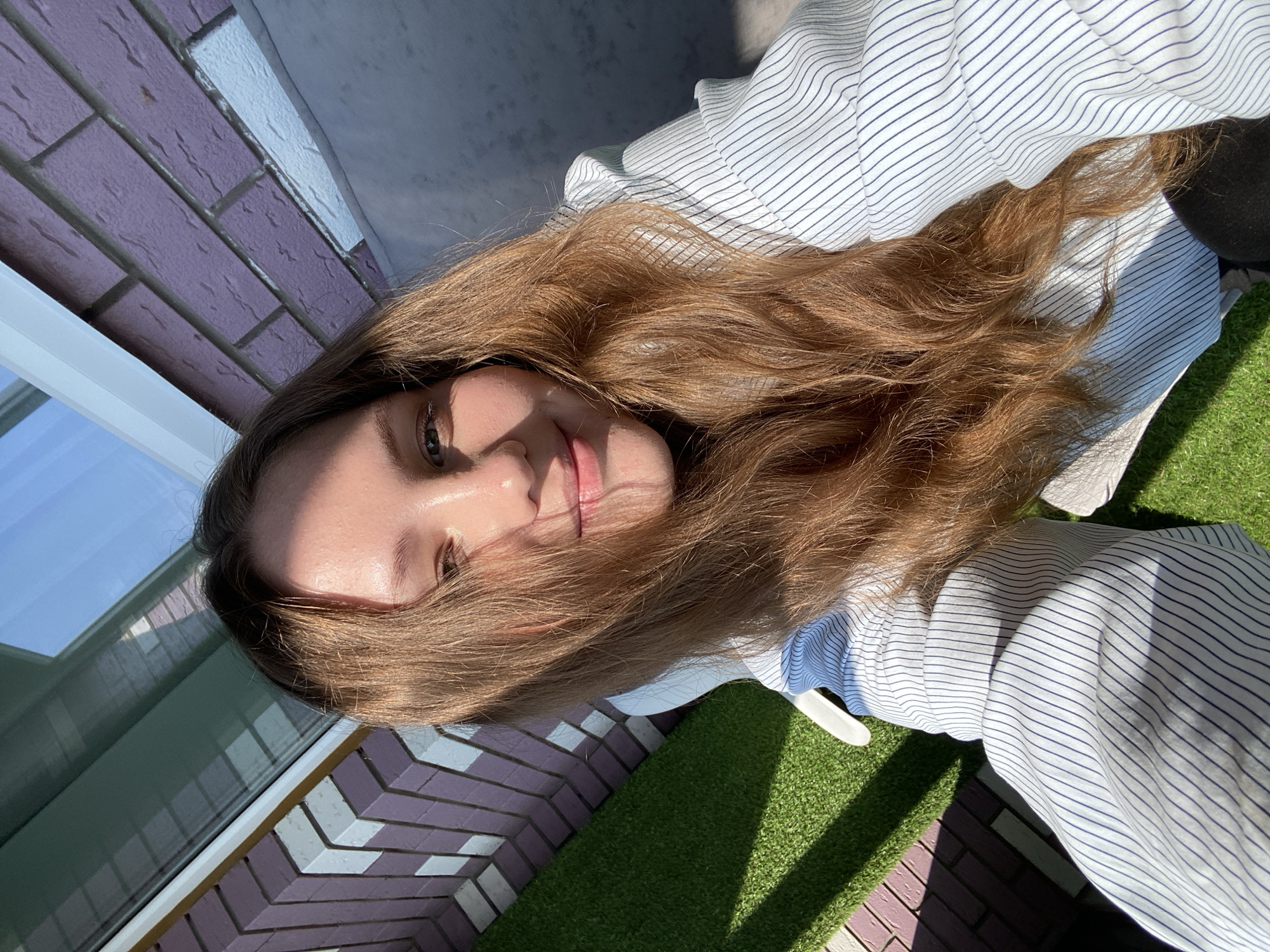 A young woman with long, light brown hair is lying down outdoors, taking a selfie. She is wearing a white and blue striped shirt. Her hair is spread out around her face and shoulders. The background shows a brick wall with a pattern of purple and white bricks, a window with a blue sky reflection, and a patch of artificial grass. Sunlight is casting shadows on the grass. The lighting suggests it is daytime. The overall mood appears relaxed and casual. The image was likely taken in Izhevsk, Russia. There is no visible text in the image.