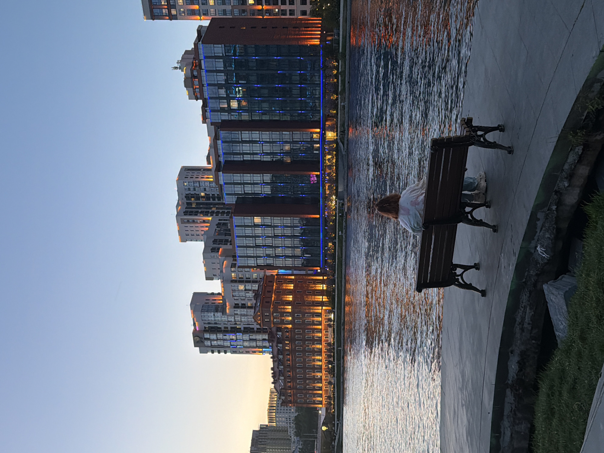 A person is sitting on a bench next to a body of water with illuminated buildings in the background, likely taken in Yekaterinburg, Russia. The buildings are varied in height and architectural style, with some displaying modern glass facades and others showcasing brickwork. The lighting suggests it is dusk or evening. The person on the bench is facing the water and buildings. The water reflects the lights from the buildings. A concrete walkway extends along the water's edge, with some greenery visible on the right.