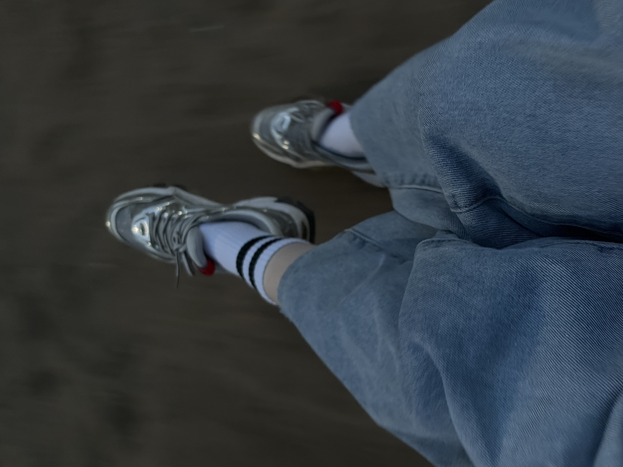 The image shows a person's legs and feet. The person is wearing light blue jeans, white socks with black stripes, and silver sneakers. The person appears to be walking on a paved surface. The location is Unknown, Unknown.