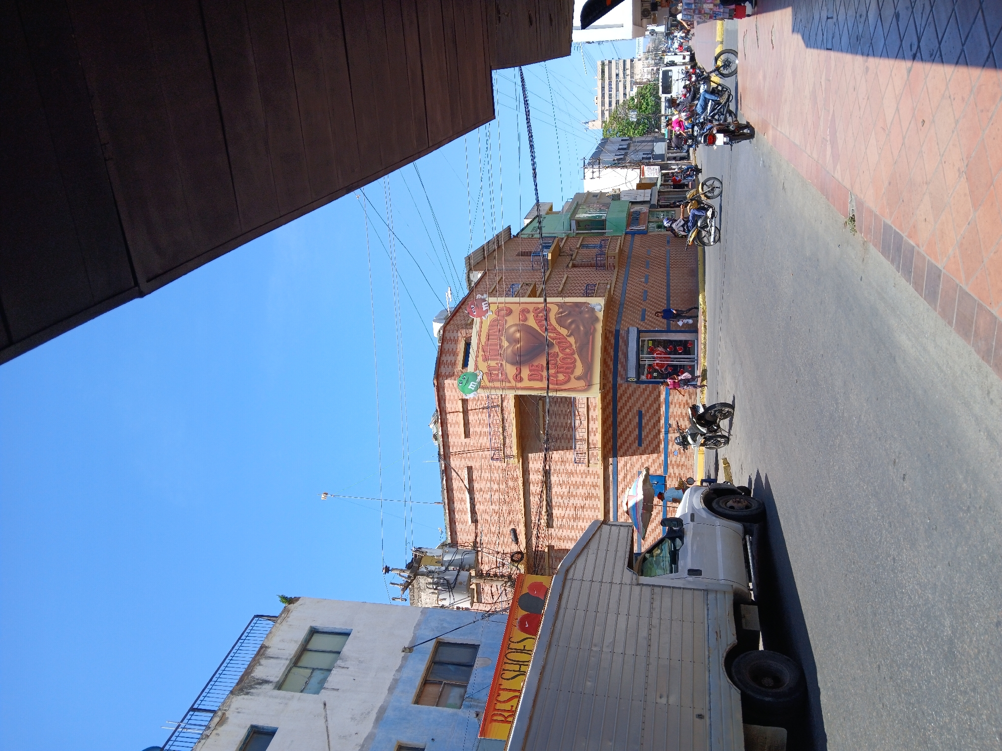 The image shows a street scene in Porlamar, Venezuela. A white box truck is partially visible on the right side of the frame, obscuring some of the view. Above the truck, a building with an advertisement for "Best Shoes" is visible. Further up, there is a multi-story building with a large mural depicting a nautilus shell and other marine-themed imagery, suggesting a seafood restaurant or related business. Several motorcycles are parked on the street. People are walking on the sidewalk in the distance. The sky is bright blue. The pavement is a mix of asphalt and patterned red brick. Overhead, there are power lines crisscrossing the scene. Part of a dark building overhangs the scene at the top left.