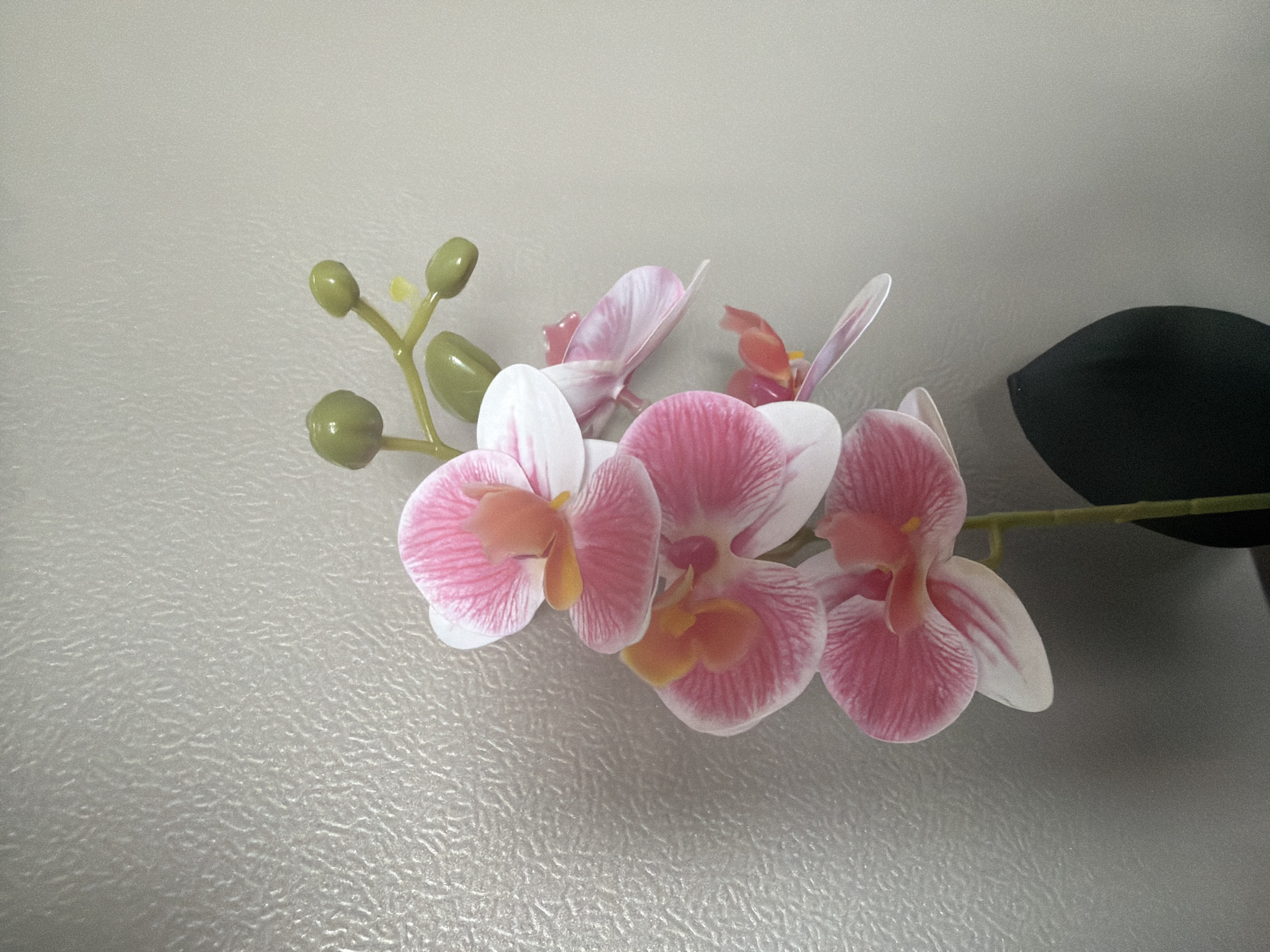 A close-up, vertically oriented shot displays a faux orchid with multiple pink and white petals, accented by an orange center. The orchid stem is a pale green, and a few small, green buds are visible at the top of the stem. The flower is set against a textured, light gray background on the left, and a plain, slightly darker gray background on the right. A dark, amorphous object, possibly a pot or a shadow, occupies the lower right corner. The lighting is subdued, creating soft shadows. This image does not contain any information that can be used to determine its location in Yuzhno-Sakhalinsk, Russia, or any other city. There are no people, actions, or interactions depicted.